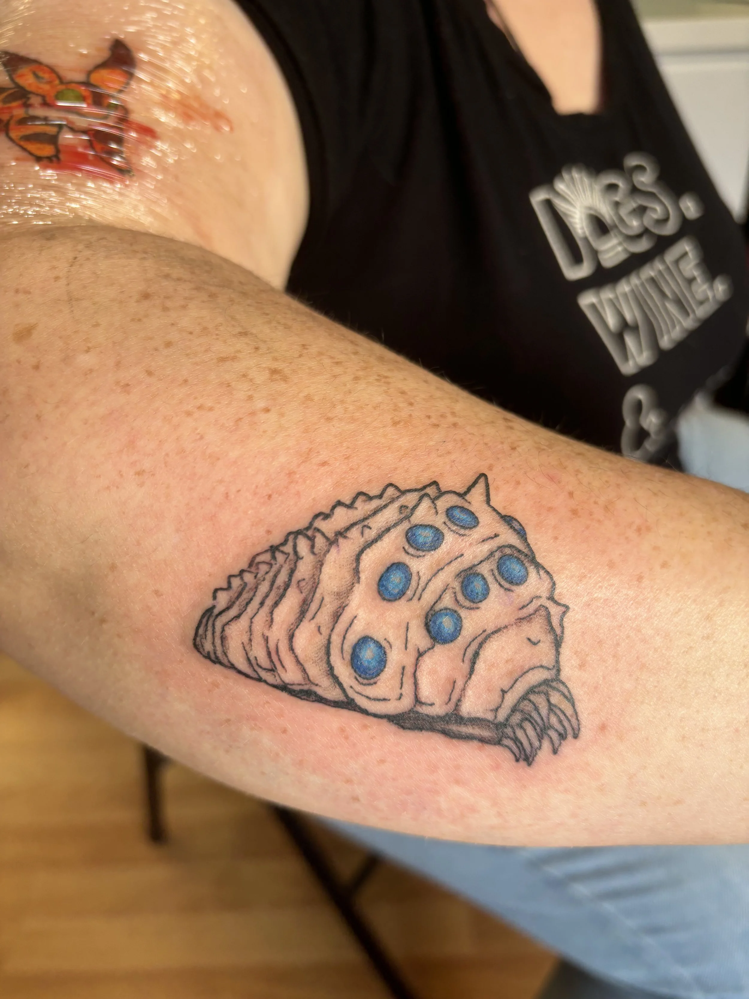Tattoo of a large, segmented, insect-like creature with multiple blue spots on its back, located on a person's upper arm.