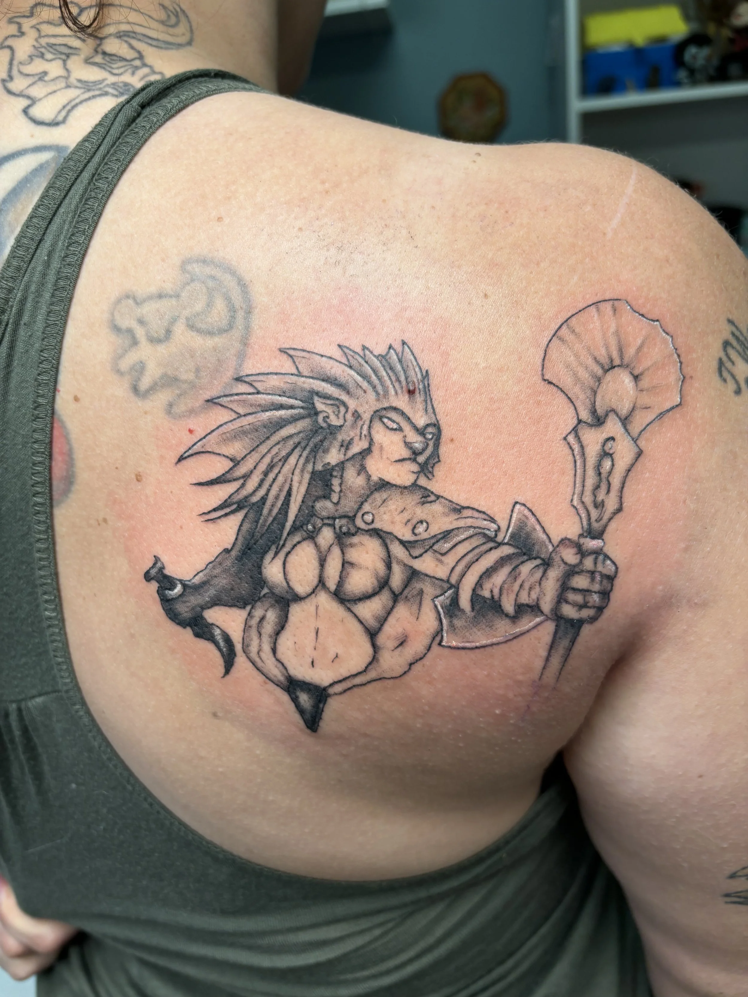 Black tattoo of a lion-headed humanoid warrior with tribal markings, holding a spear, on a person's shoulder.