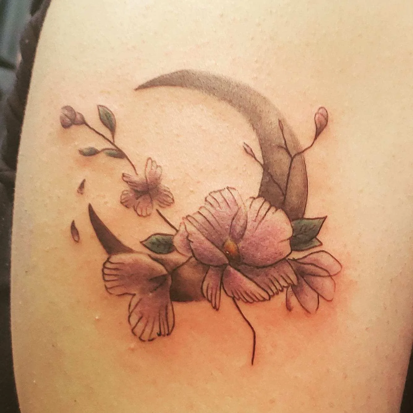 Tattoo of a crescent moon with pink flowers and green leaves on skin.