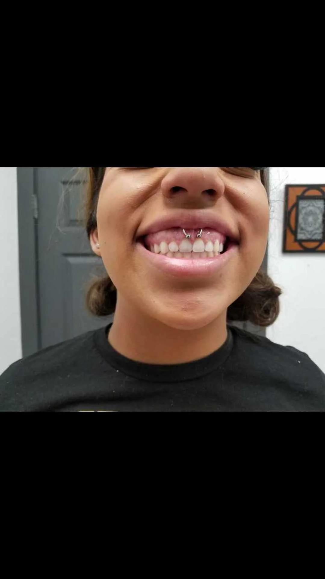 Person smiling with silver septum piercing showing, wearing a black shirt indoors.