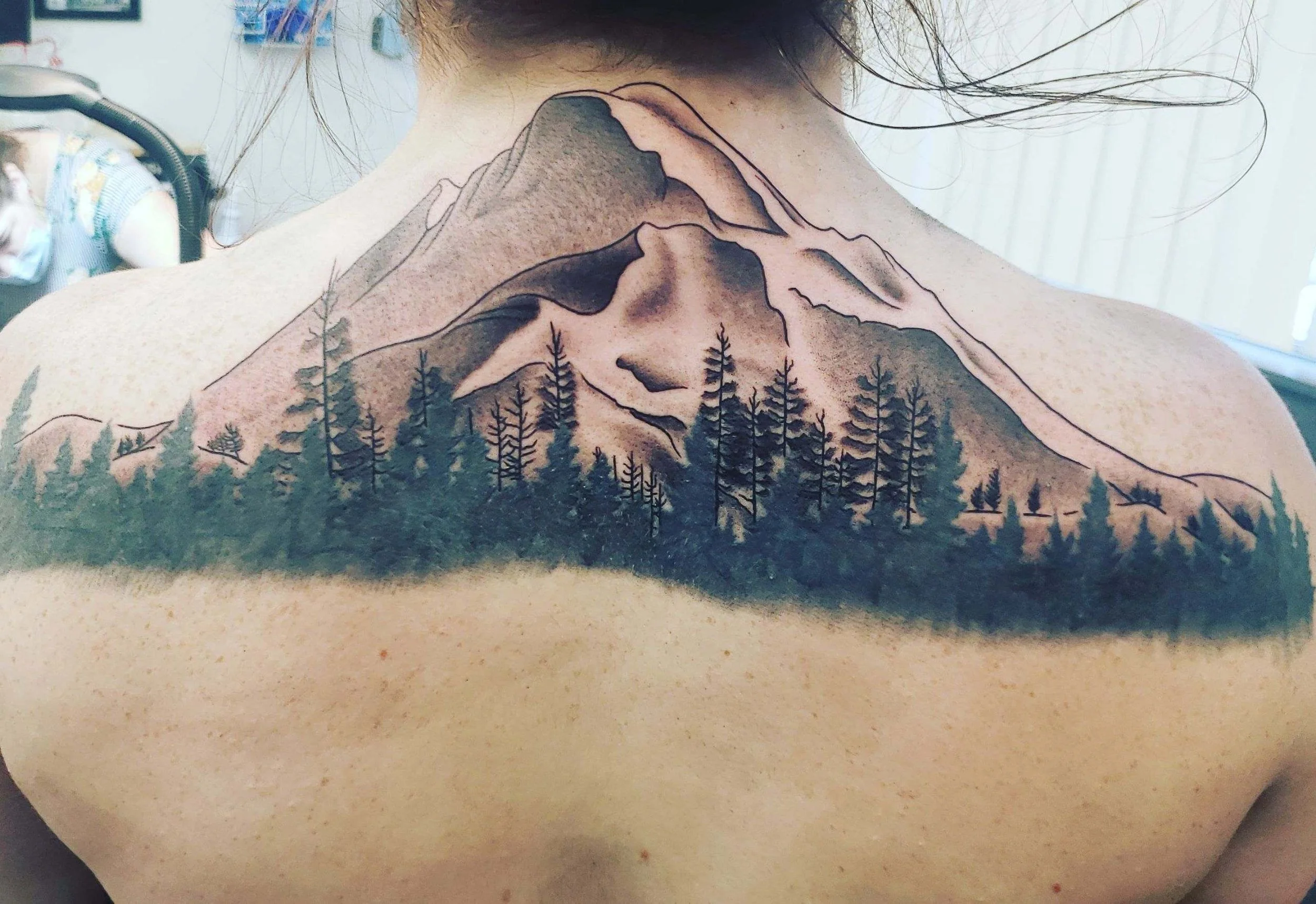 Detailed tattoo of a mountain landscape with pine trees on a person's upper back.