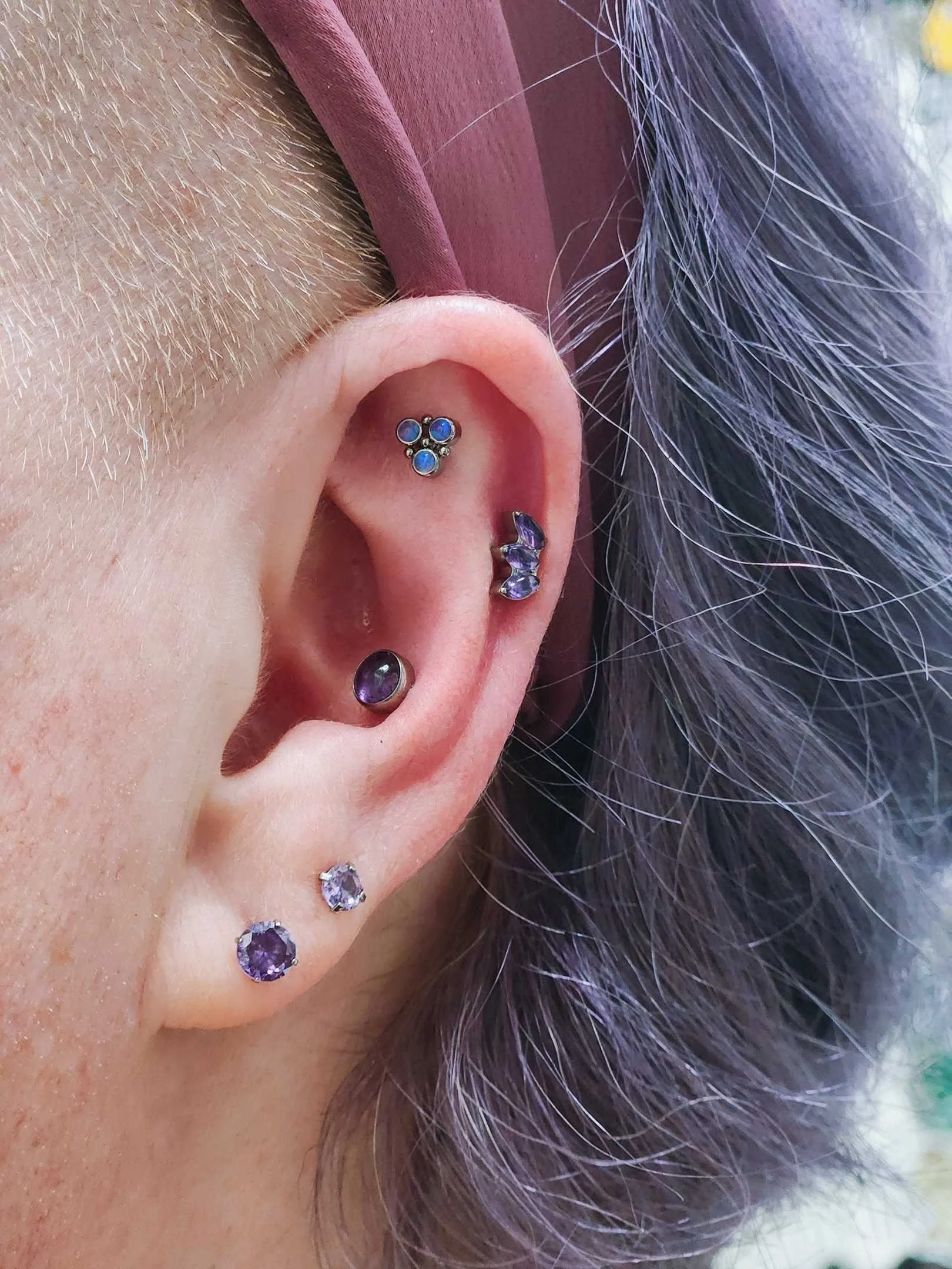 Close-up of an ear with multiple piercings, featuring various gemstone and metal stud earrings, with purple hair partially visible.
