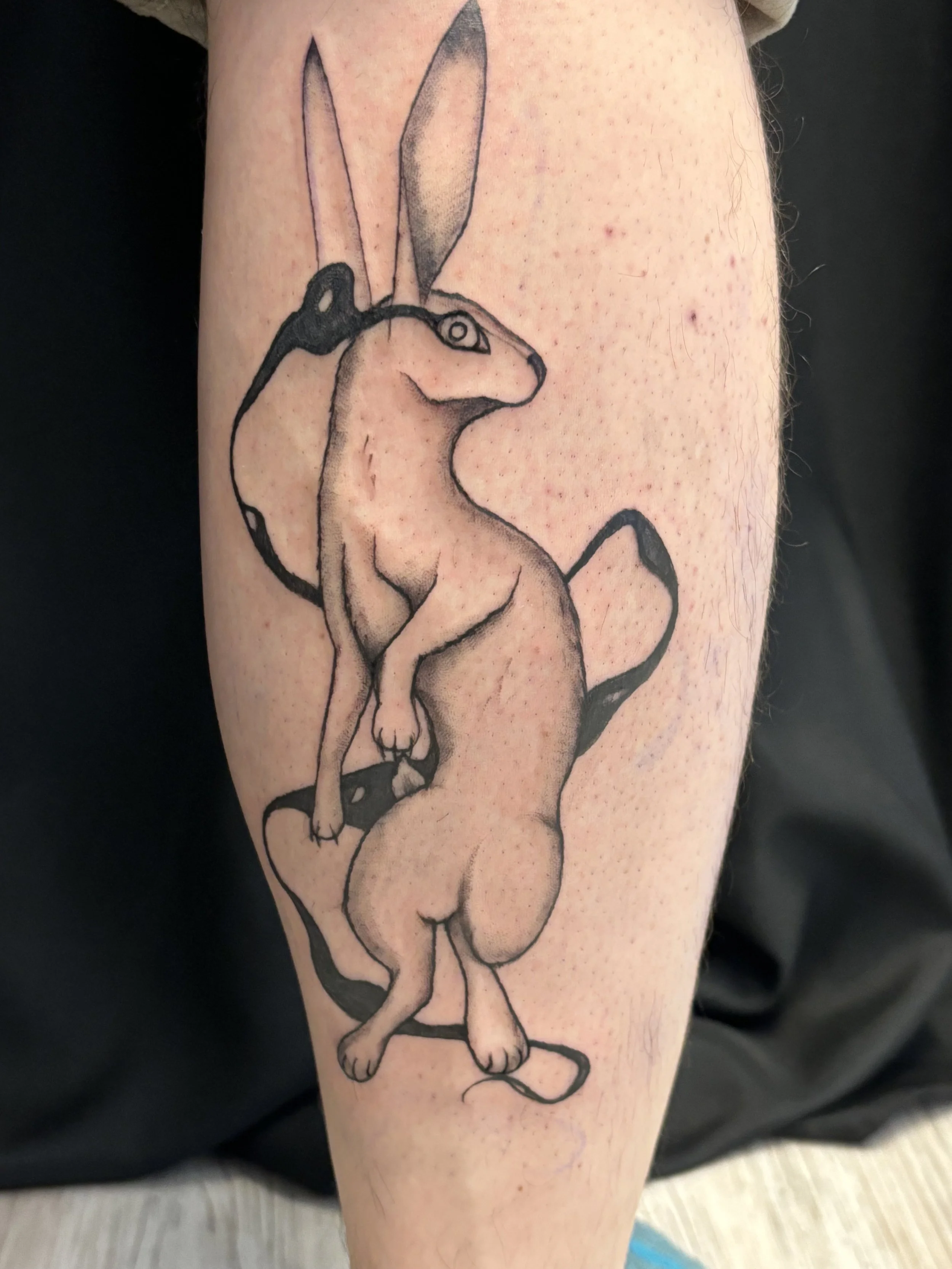 Tattoo of a mythical creature blending a rabbit and a dog, with a long body, large hindquarters, and a tail, drawn with bold black lines and shading.