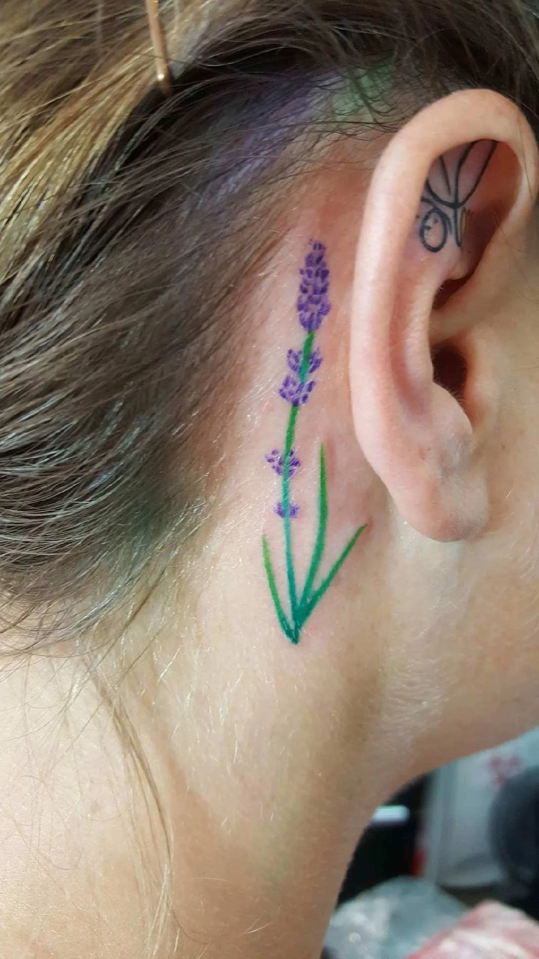 Close-up of a person's ear with a small tattoo of a purple lavender flower behind it.