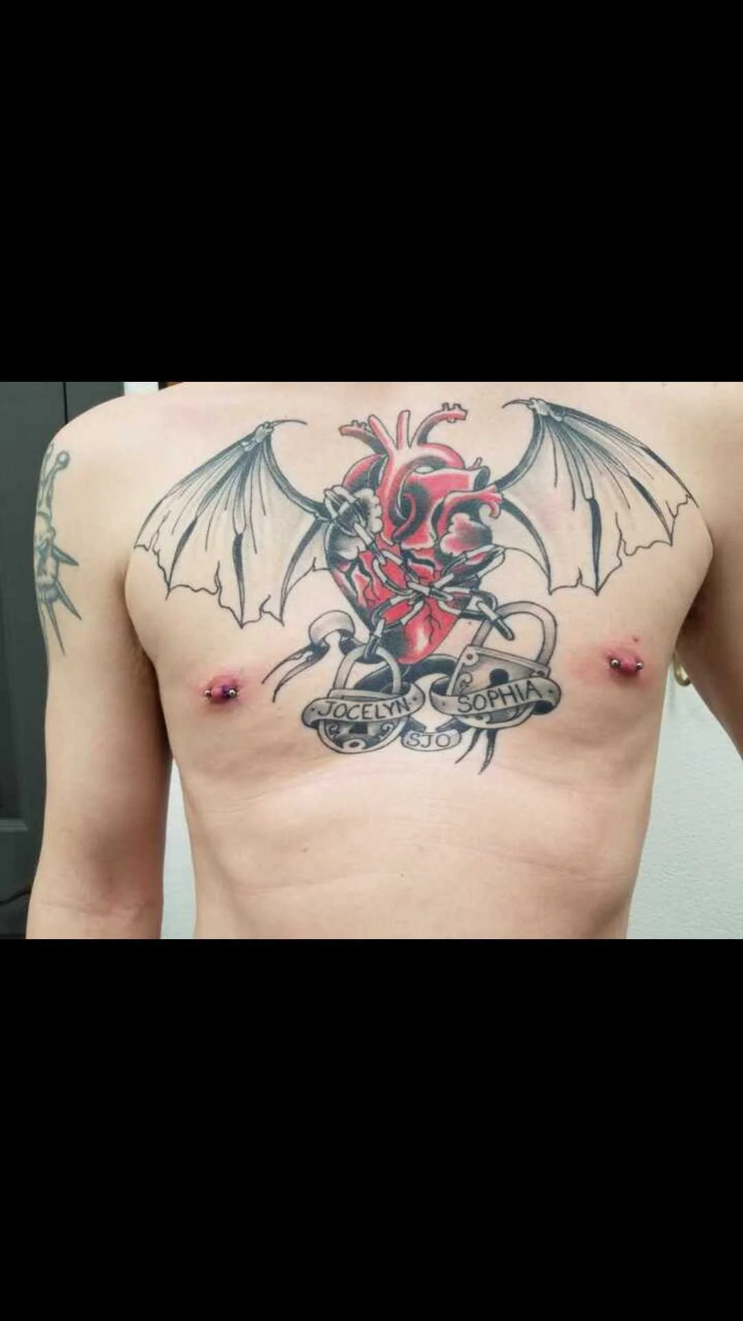 Chest tattoo featuring a stylized red human heart with bat-like wings and two ribbon banners. The ribbons have text and are decorated with heart lock designs. The person also has nipple piercings.