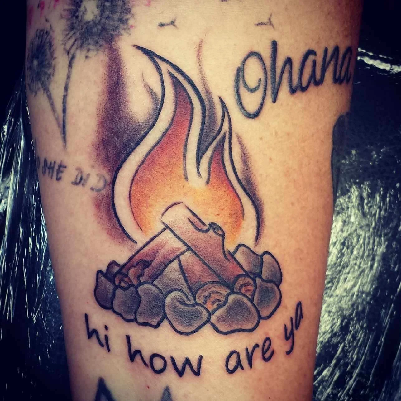 Tattoo of a campfire surrounded by stones with the words "hi how are ya" below and "Ohana" above, along with dandelion images.