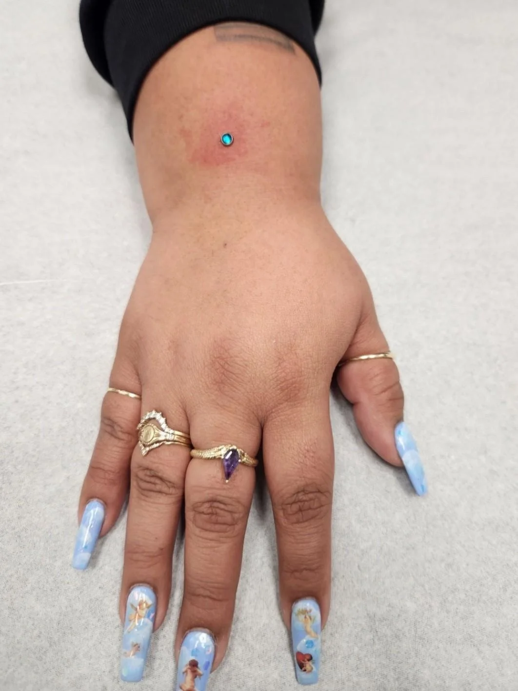 A hand with blue-themed nail art featuring decorative designs. The person is wearing gold rings, one with a purple stone. There is a small piercing with a turquoise gem on the upper part of the hand.