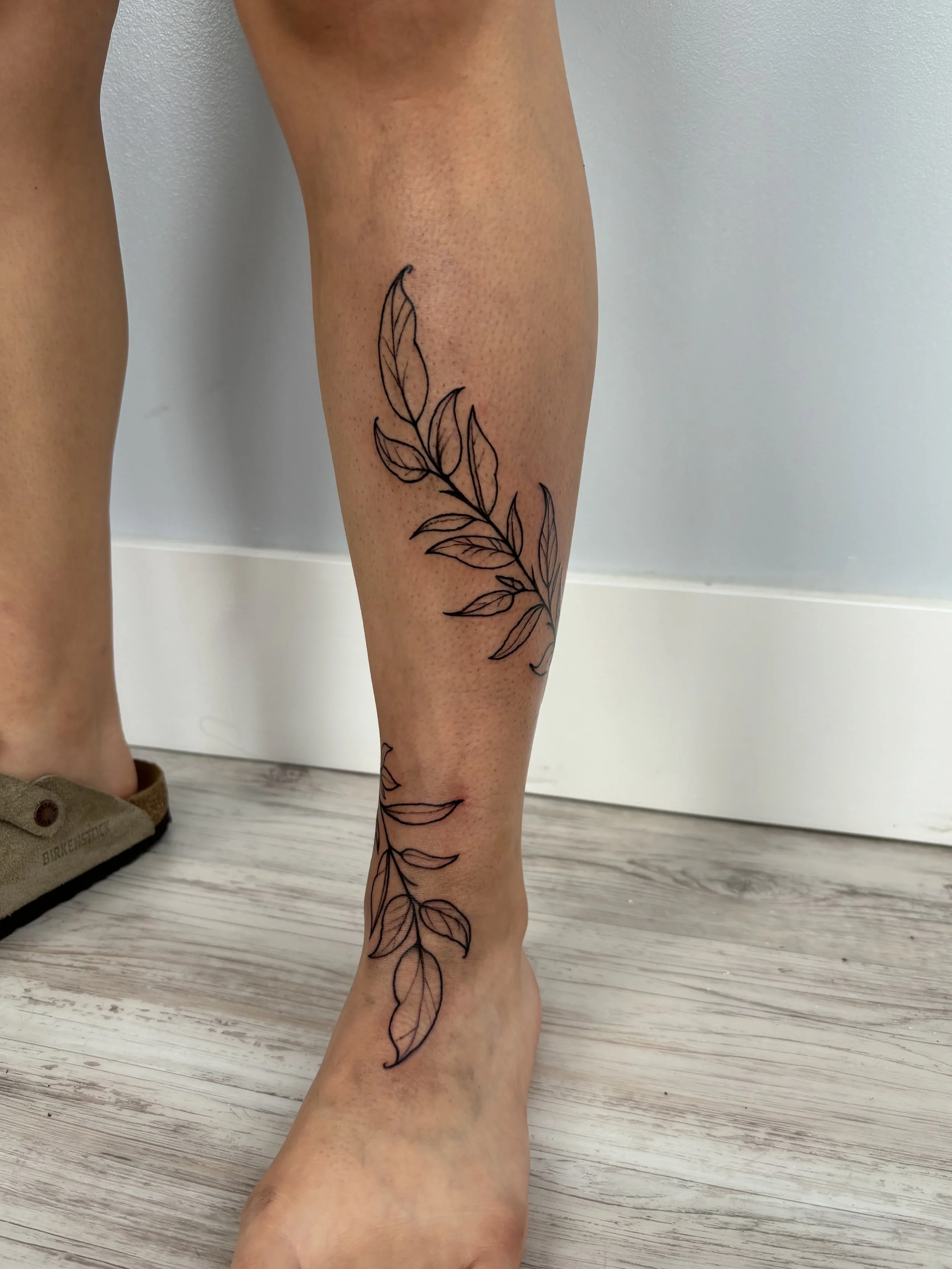 A person's lower leg with a botanical tattoo of leaves and branches in black ink.