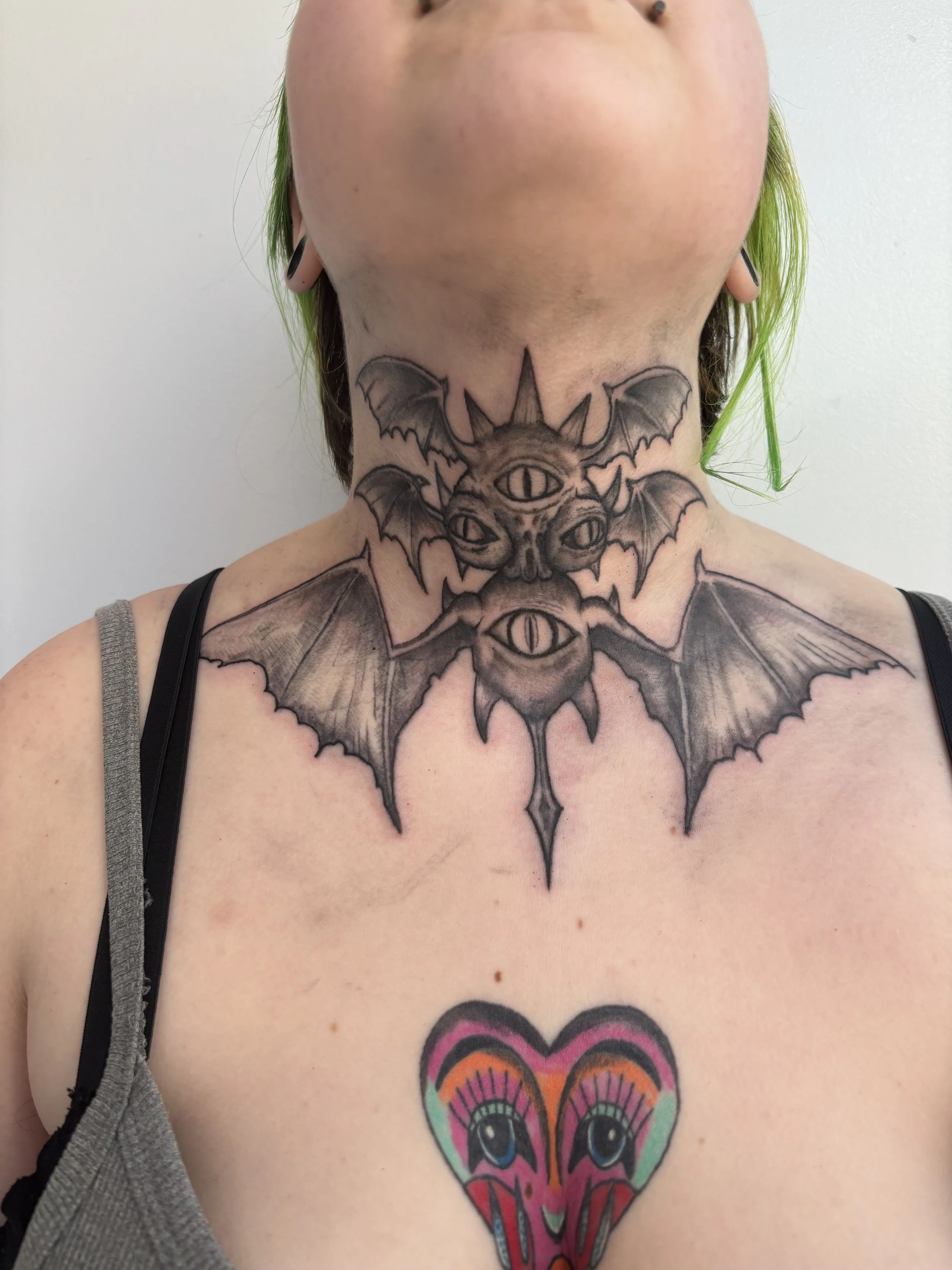 Tattoo of a bat-like creature with three eyes and wings on a person's neck with colorful heart-shaped design on front of chest.