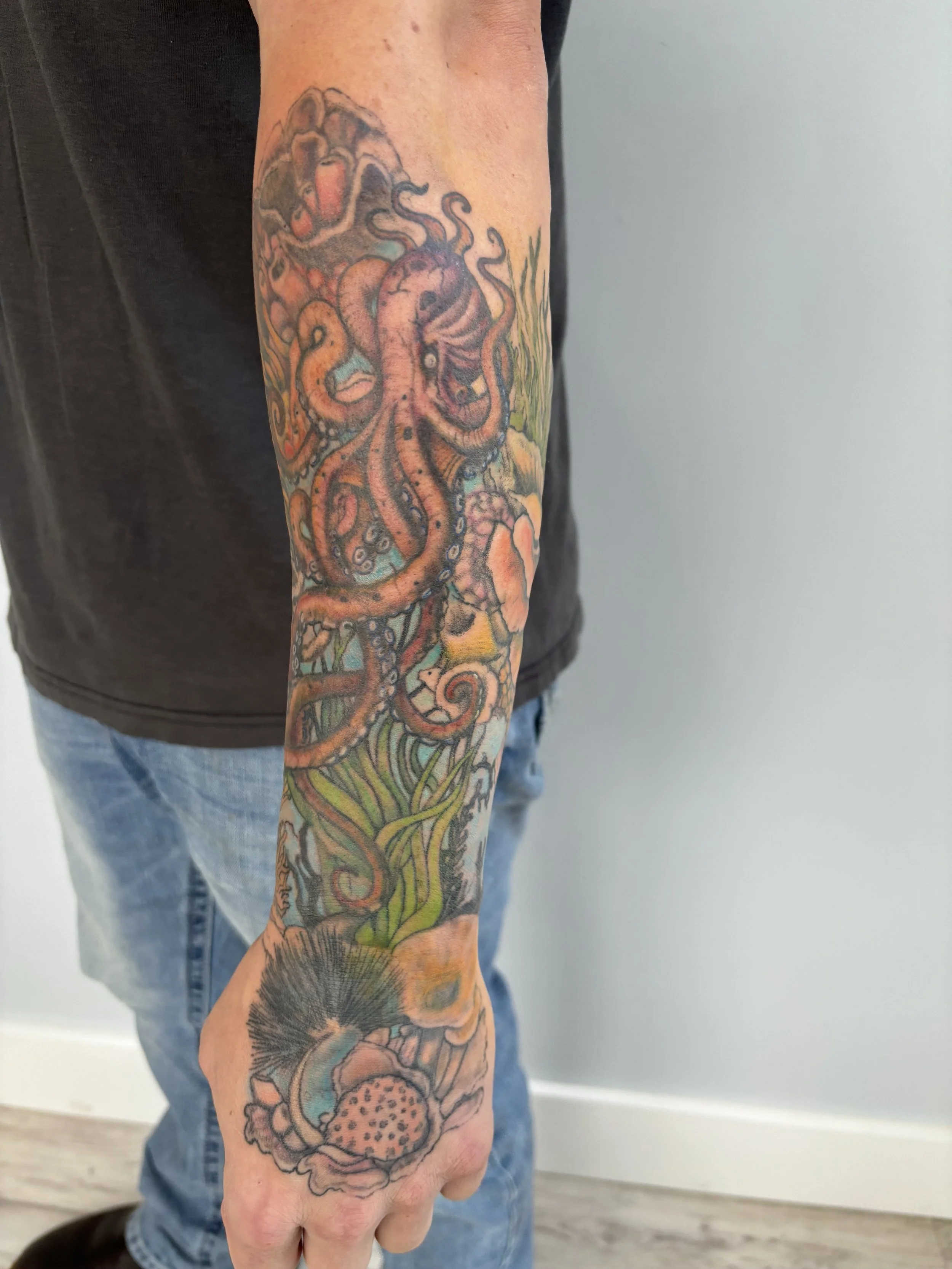 Colorful tattoo of an octopus with tentacles wrapping around the forearm, with coral and seaweed at the base.