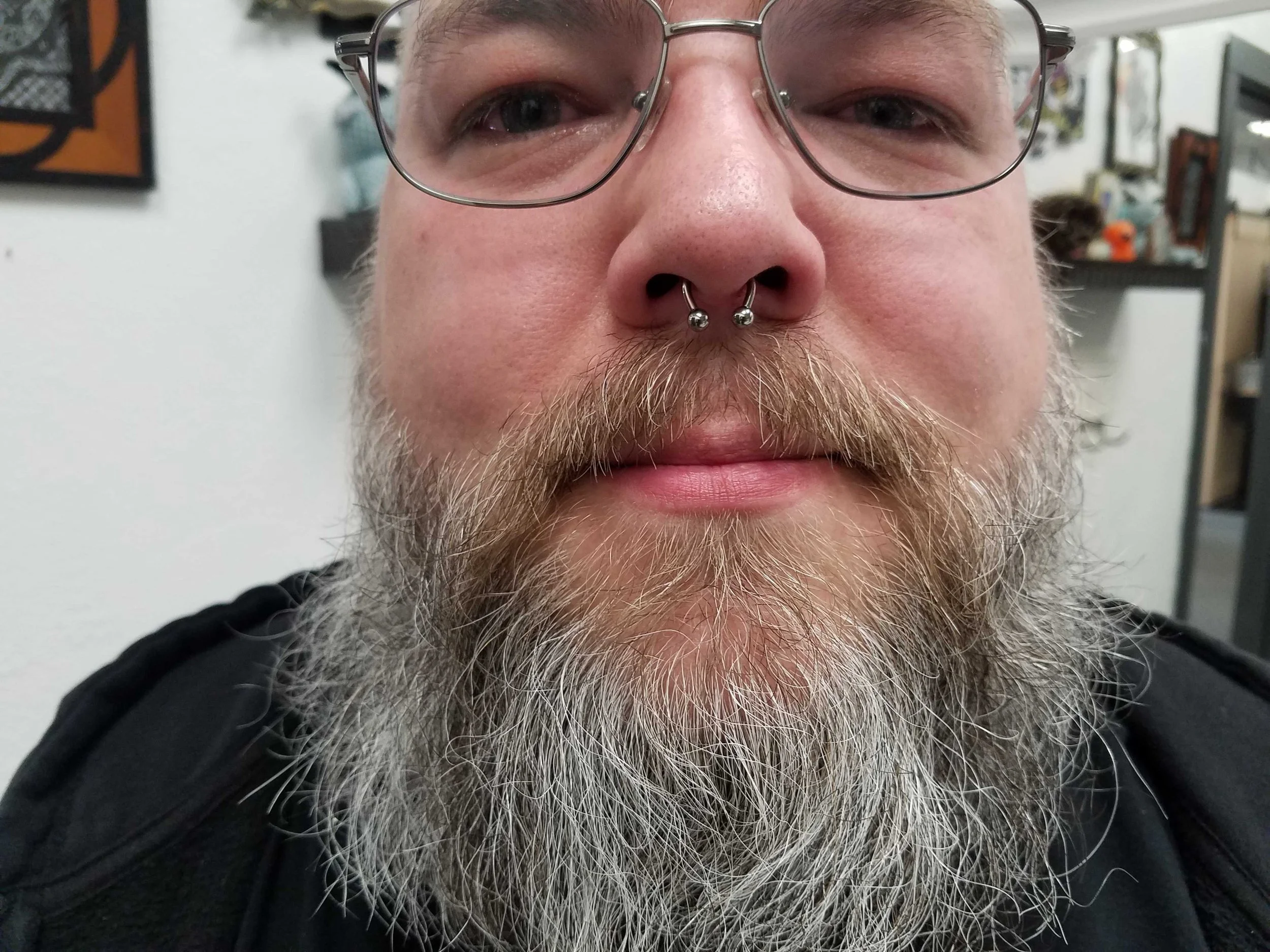 Close-up of a person with a gray beard and glasses, wearing a septum piercing.