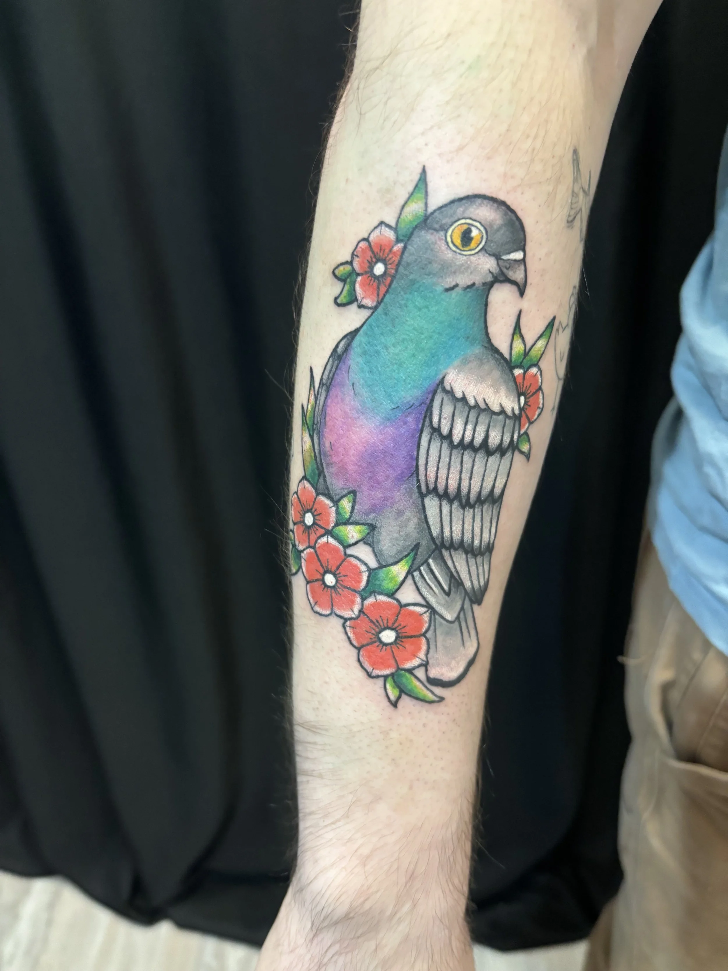 Colorful tattoo of a bird with vibrant feathers, surrounded by red flowers and green leaves.