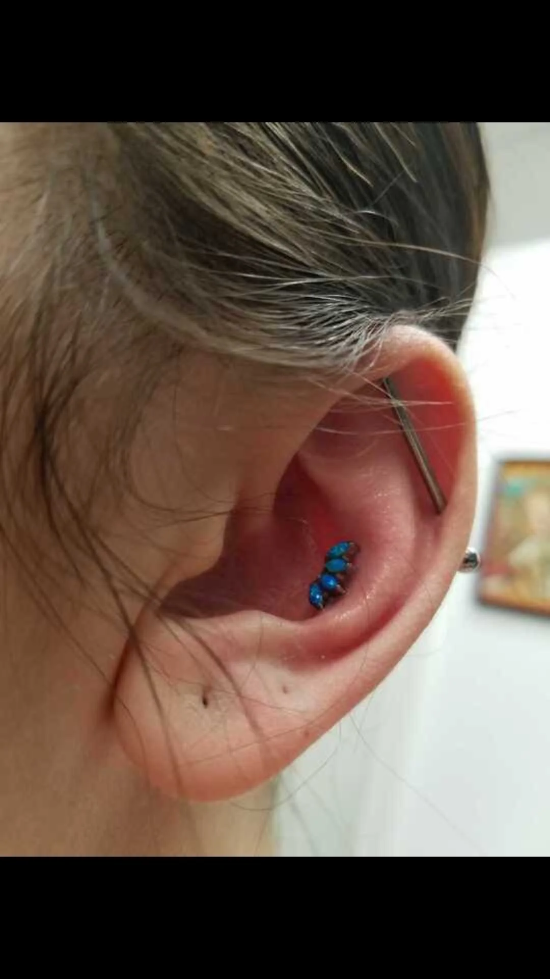Close-up of an ear with multiple piercings, including a blue gemstone conch piercing and an industrial barbell.