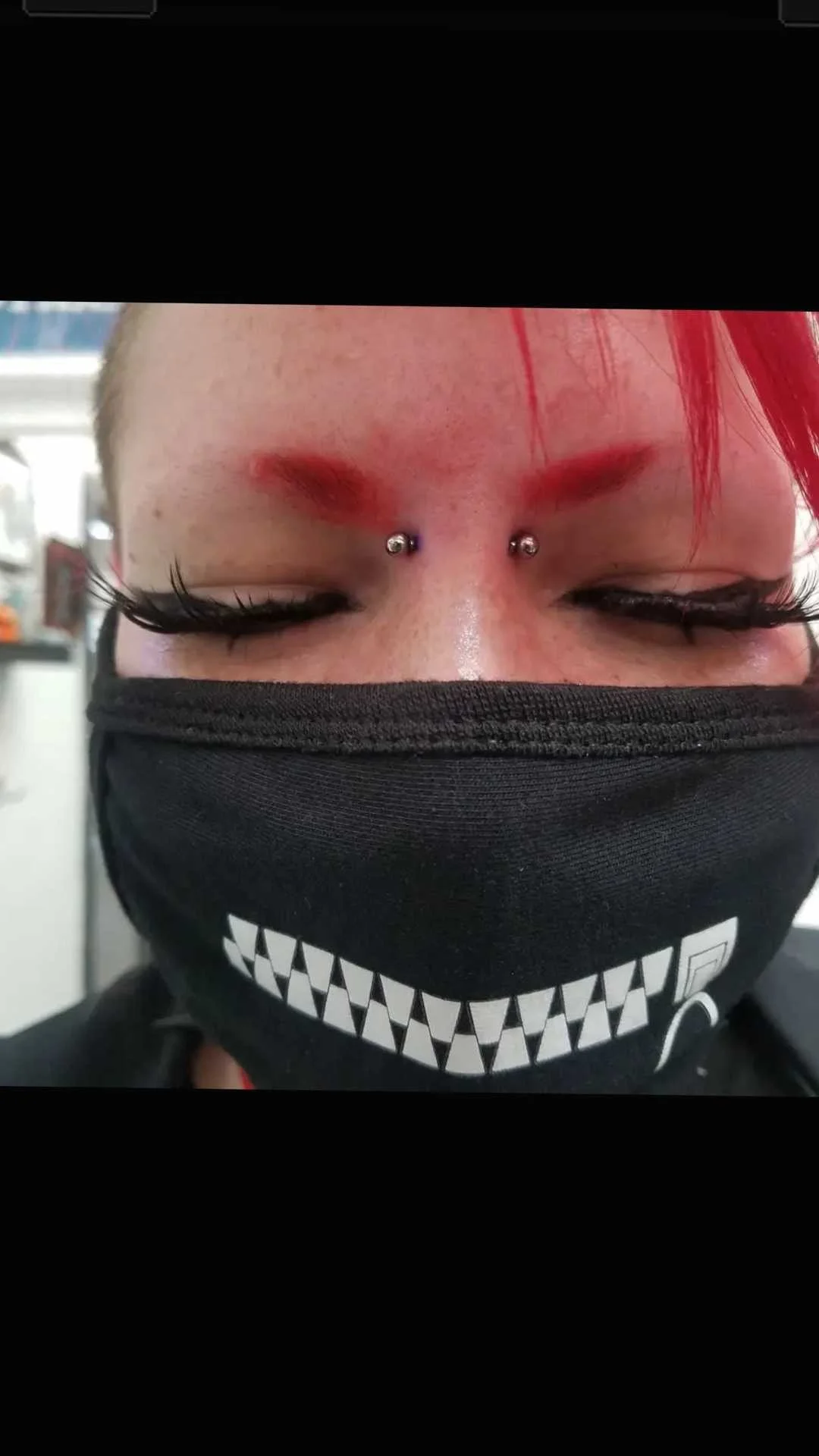 Close-up of a person with bright red eyebrows, eyebrow piercings, long black eyelashes, and wearing a black face mask with a stylized white grin design.