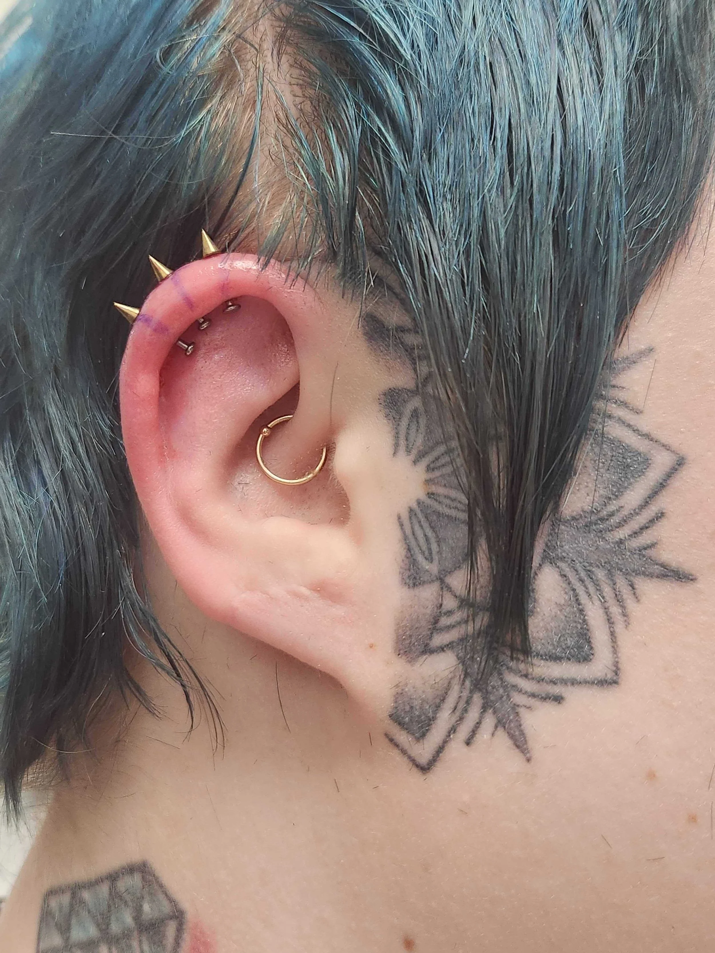 Close-up of an ear with multiple piercings and tattoos on the side of the face, including a spiked piercing on the upper ear and a gold hoop in the lower ear.