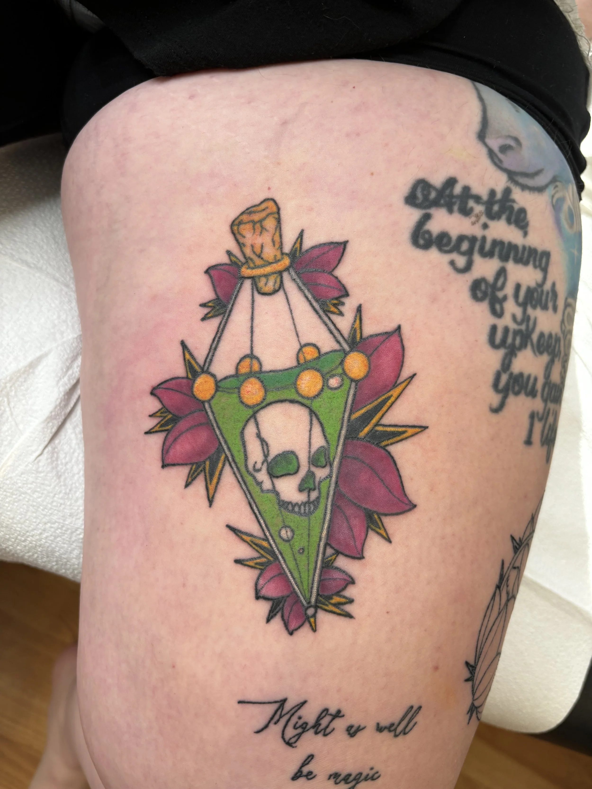 Tattoo of a skull inside a green triangle, surrounded by pink leaves and gold spikes, with a small thumbtack at the top connected to a string of yellow dots.