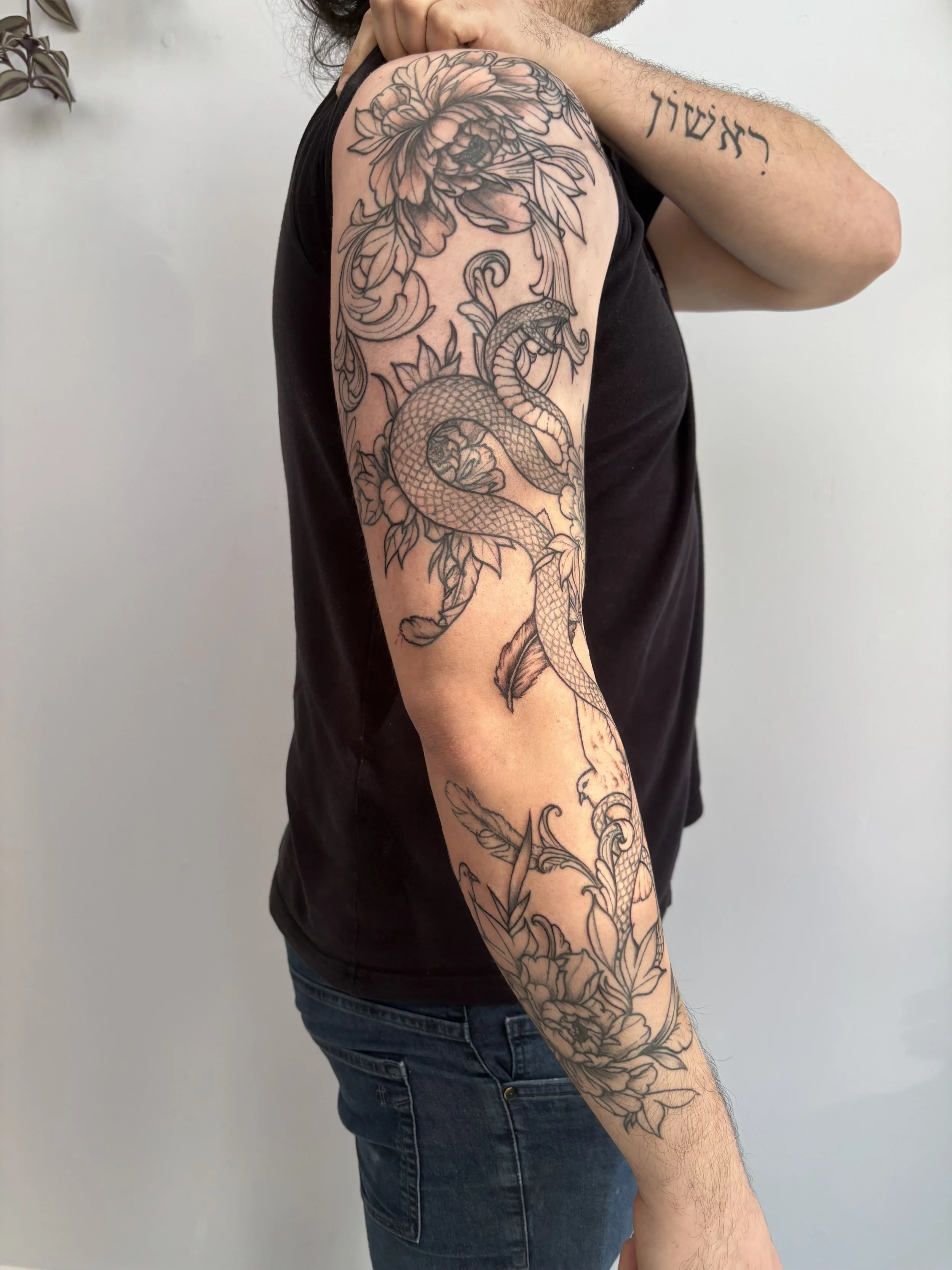 A person with a large tattoo sleeve on their upper arm and forearm featuring roses, a snake, and leaves, dressed in a black sleeveless shirt and jeans.