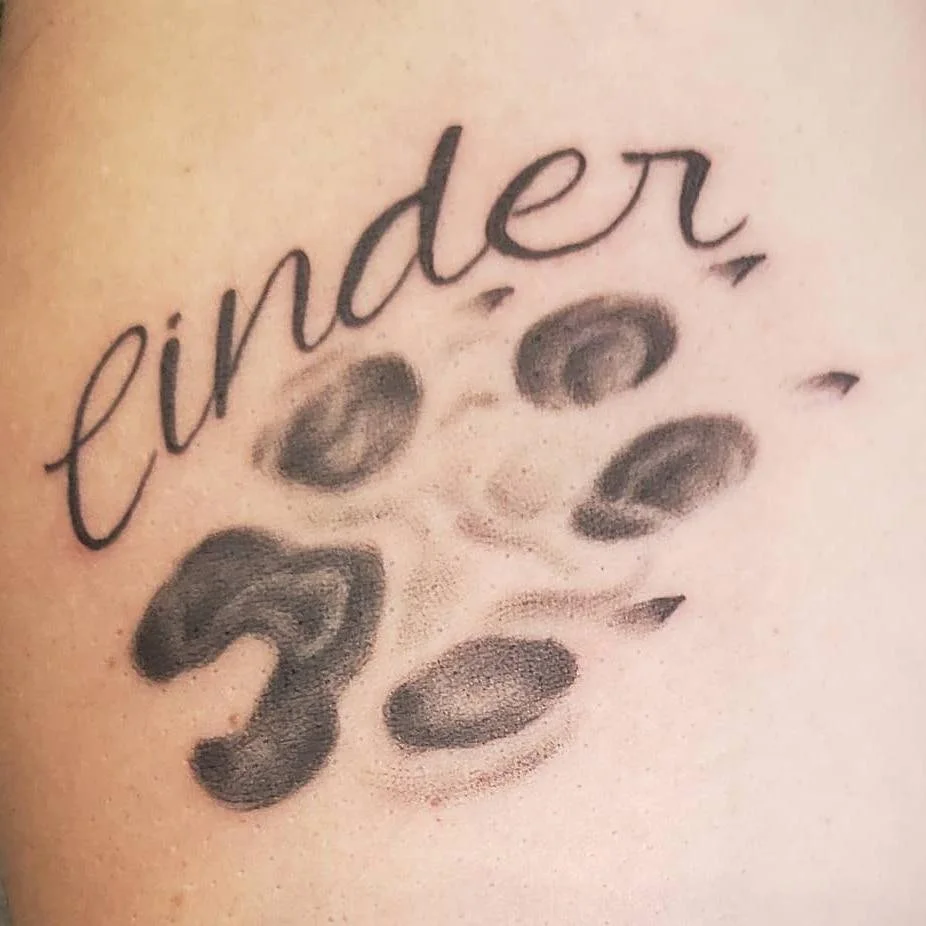 Tattoo of a paw print with the word "Cinder" in cursive script.