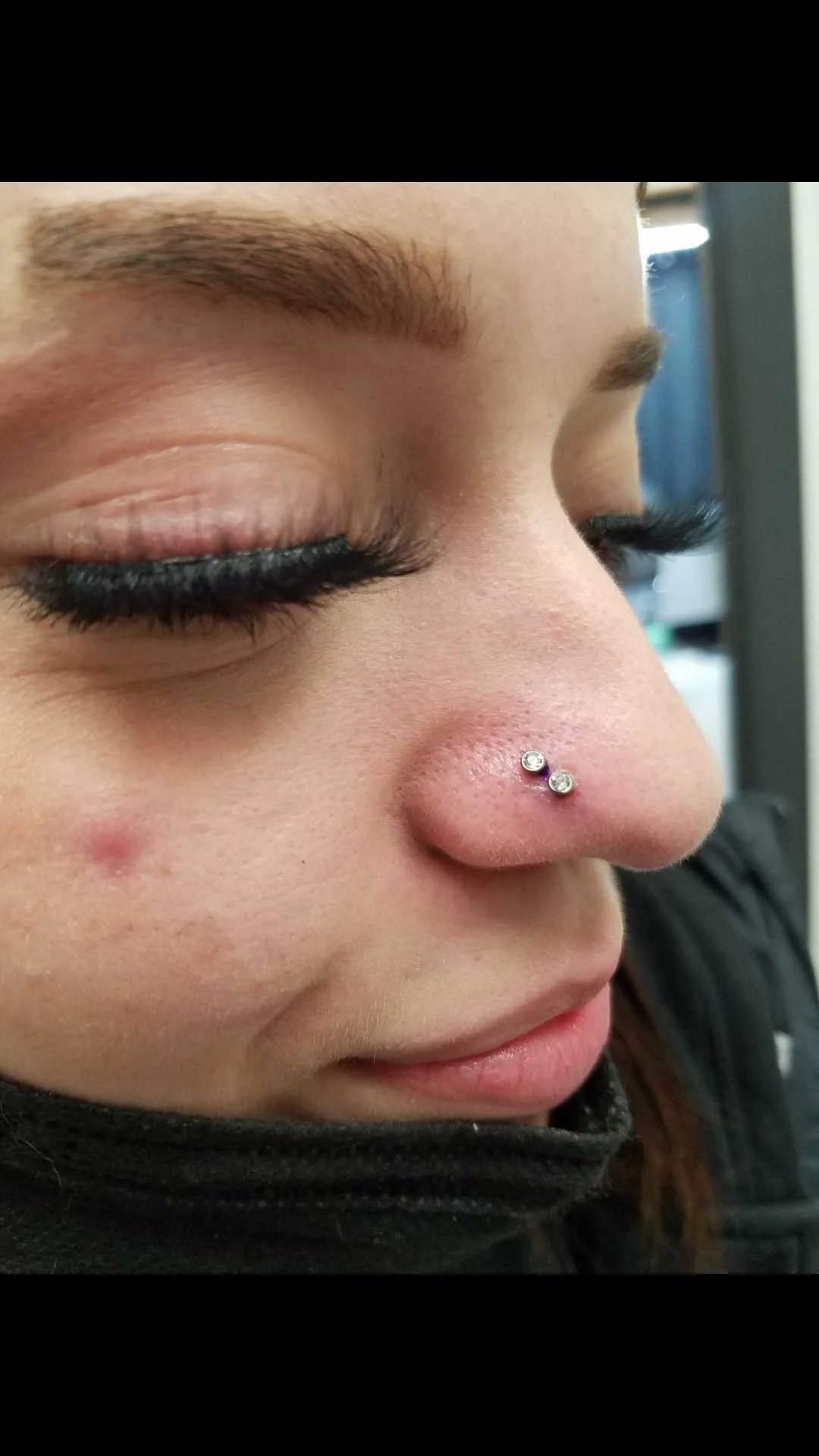 Close-up of a person's nose and cheek with a nose piercing featuring two gems, long eyelashes, and eyebrow visible.