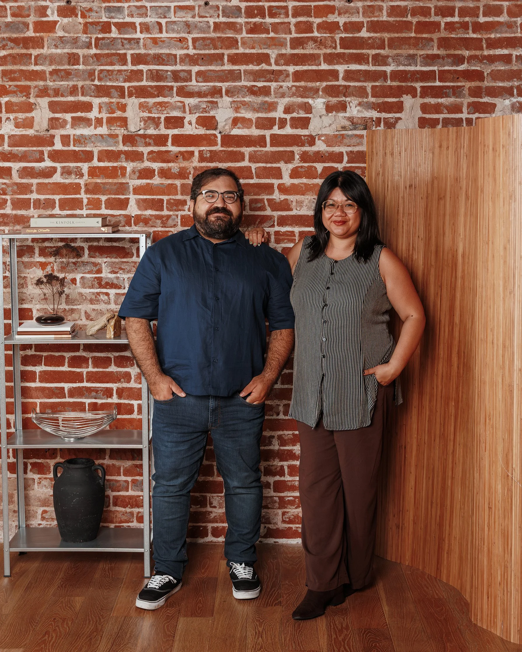 A man and woman standing together indoors, posing for a photo against a red brick wall. The man has dark hair, glasses, a beard, and is wearing a long-sleeve dark blue shirt, jeans, and sneakers. The woman has straight black hair, glasses, and is wearing a sleeveless black-and-white striped top and brown pants. They are smiling, with the woman resting her hand on the man's shoulder.