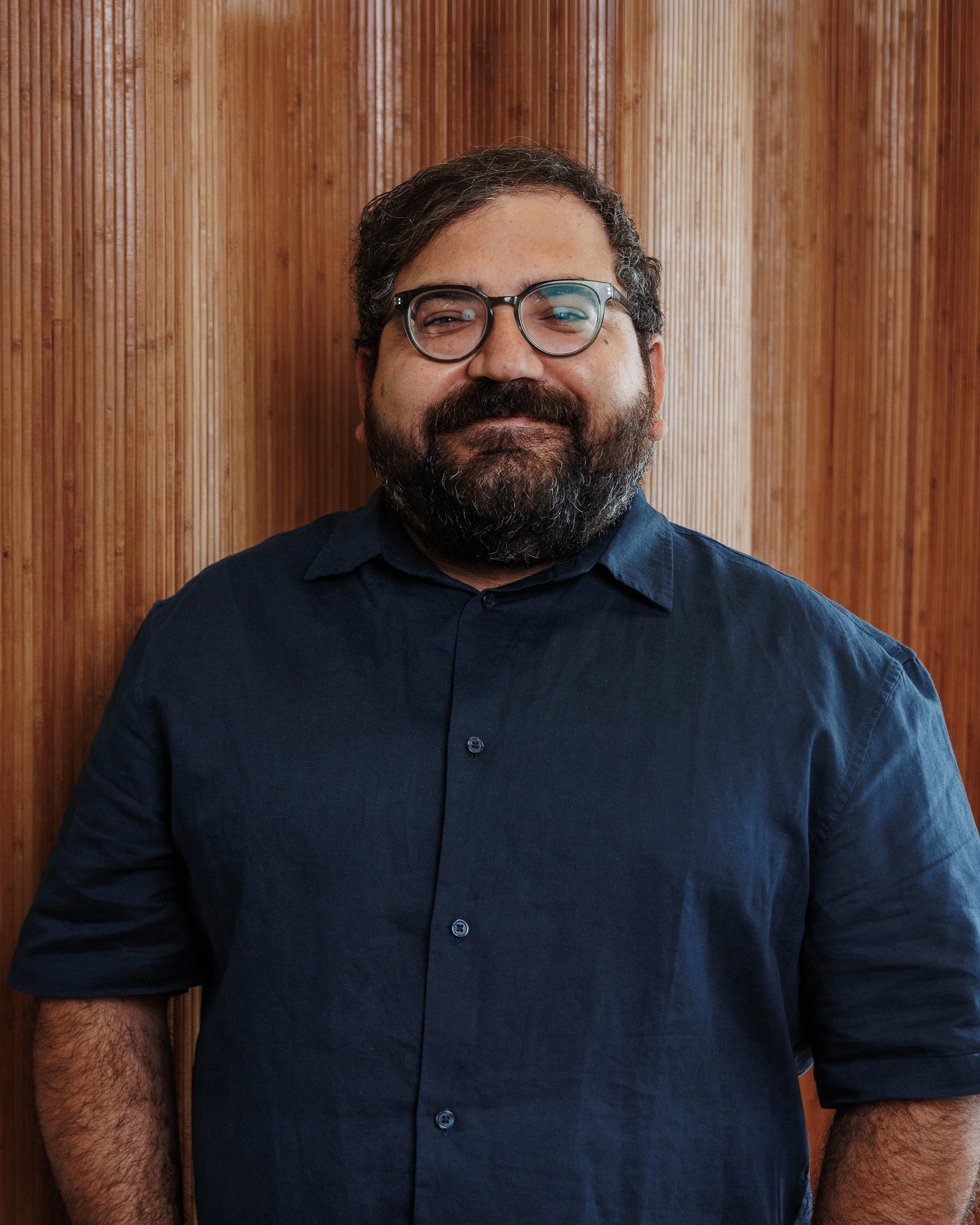Portrait of a man with glasses, a beard, and curly hair, wearing a dark green shirt and beige jacket in front of a brown background.