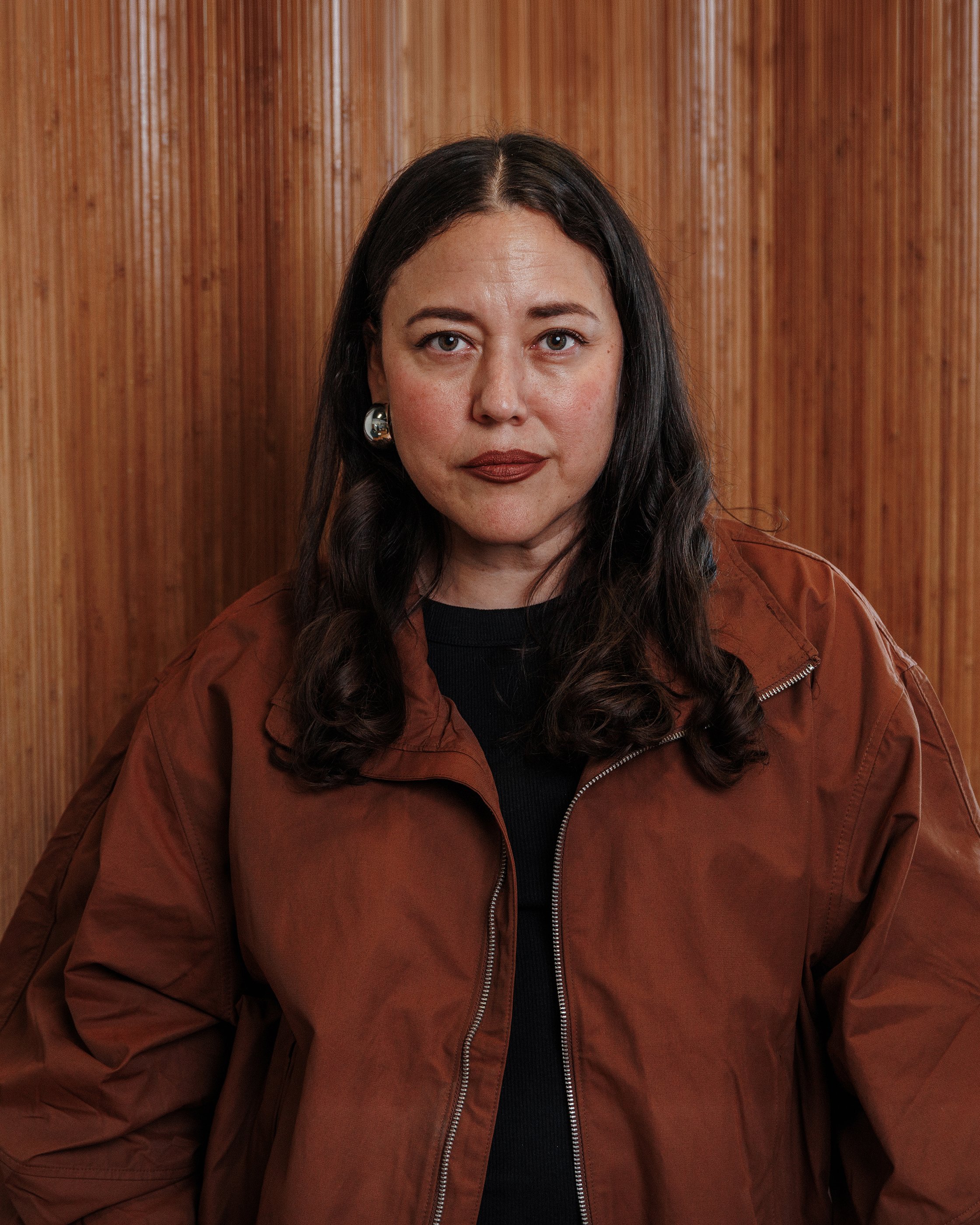 A woman with long, dark wavy hair and fair skin, wearing a plaid blazer, a black top, and a heart-shaped necklace, against a brown background.
