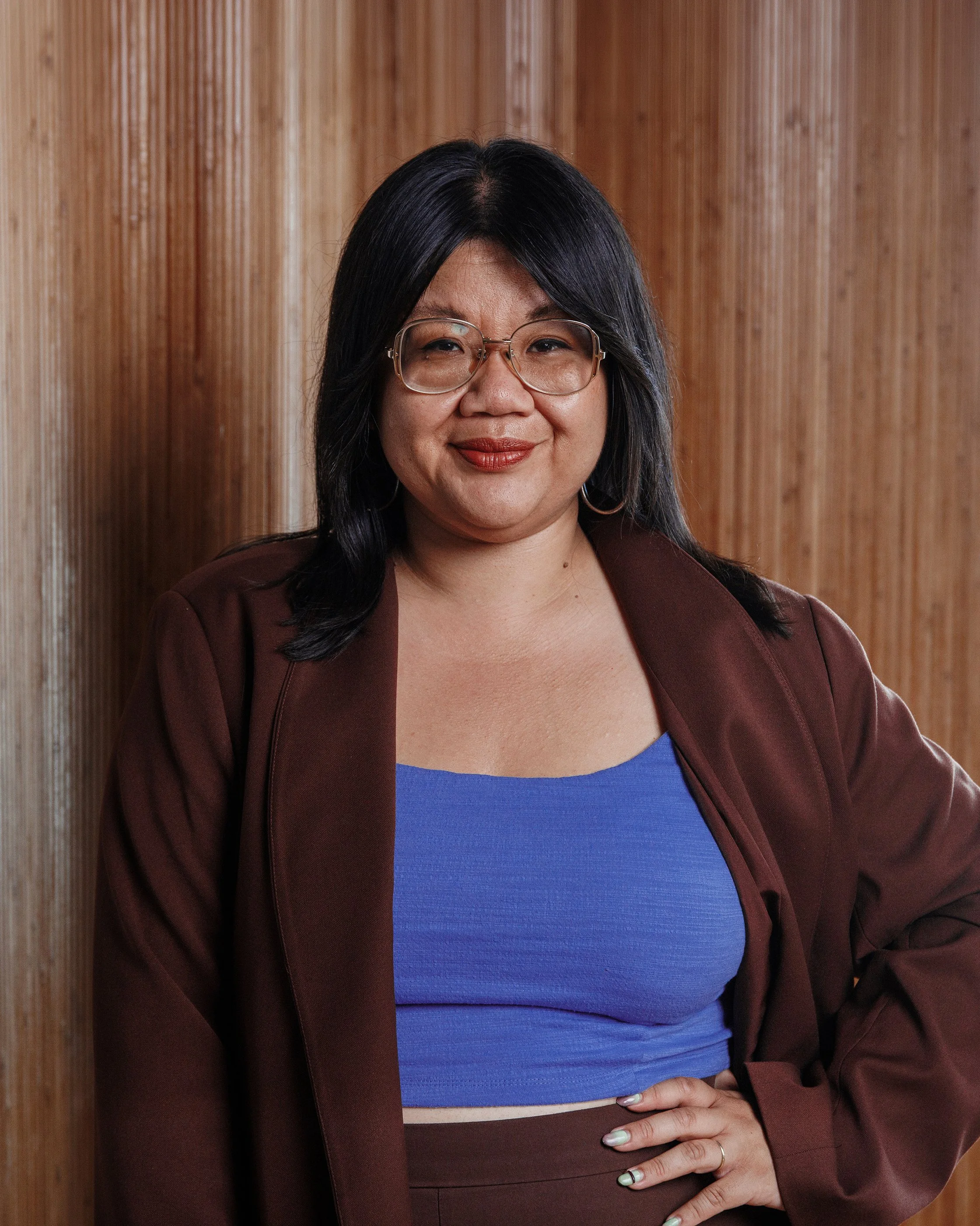 A woman with long black hair, wearing a tan button-up shirt with a decorative zipper and rings, smiling against a brown background.