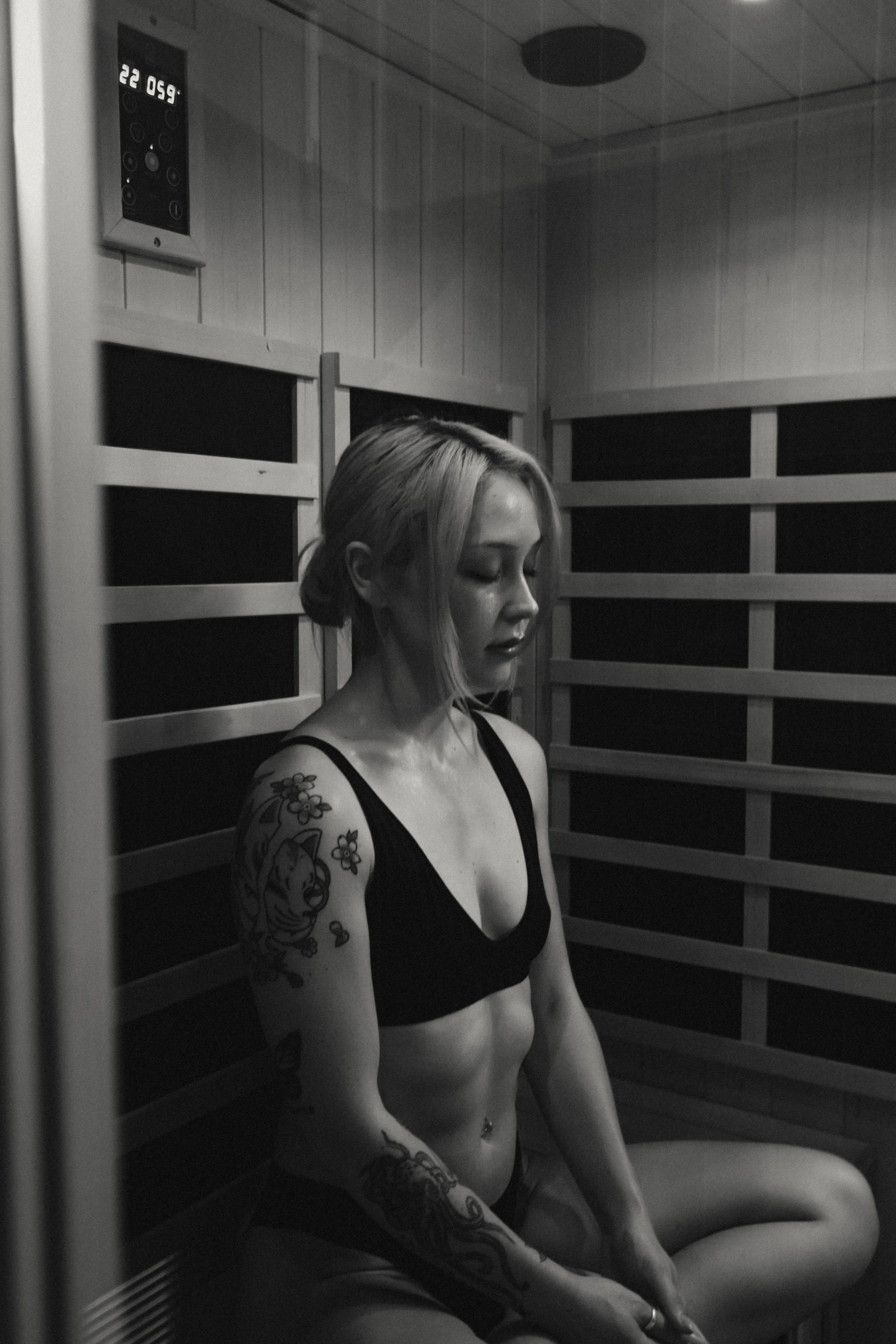 A woman with tattoos on her arm and torso sits in a sauna with wooden walls, wearing a black sports bra and shorts, with closed eyes and a serene expression.