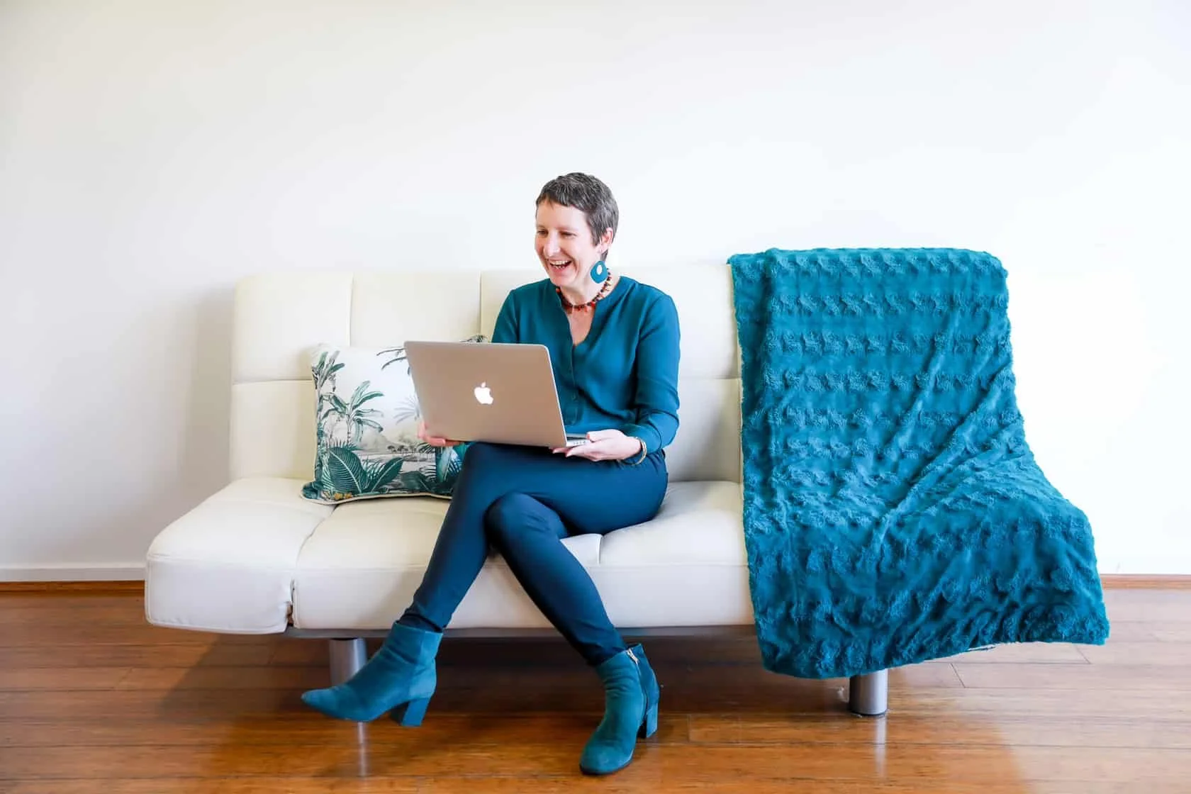 Gina is sitting on a white couch looking at meterial on a laptop.