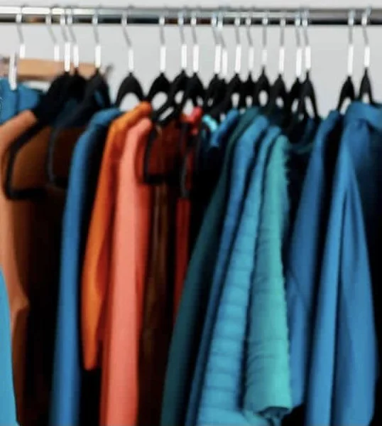 Clothes hanging on a rack, including tops, trousers and jackets in blue, teal, orange, and brown.