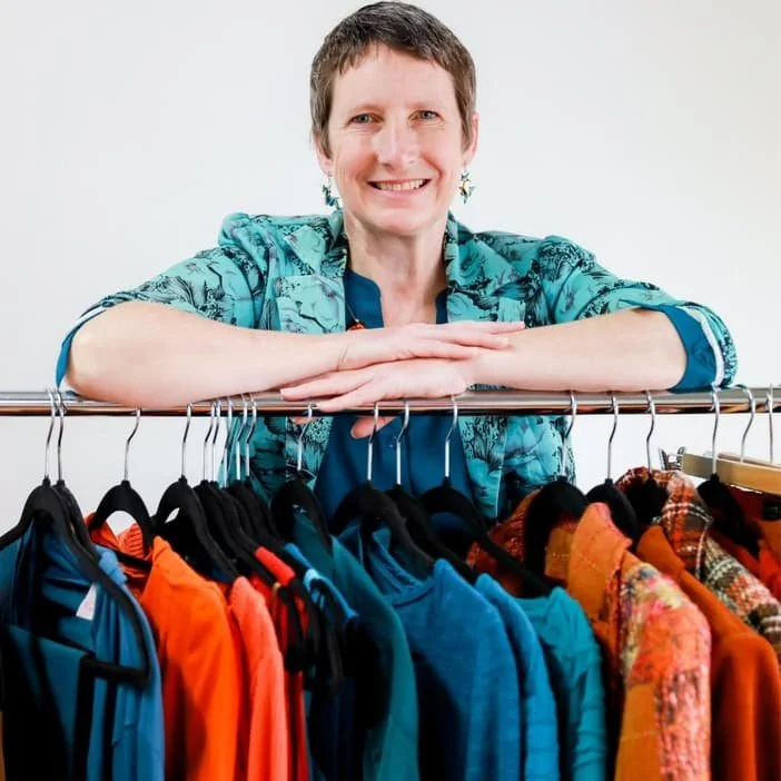 Woman leaning on a clothing rack filled with colorful jackets and shirts, smiling at the camera.