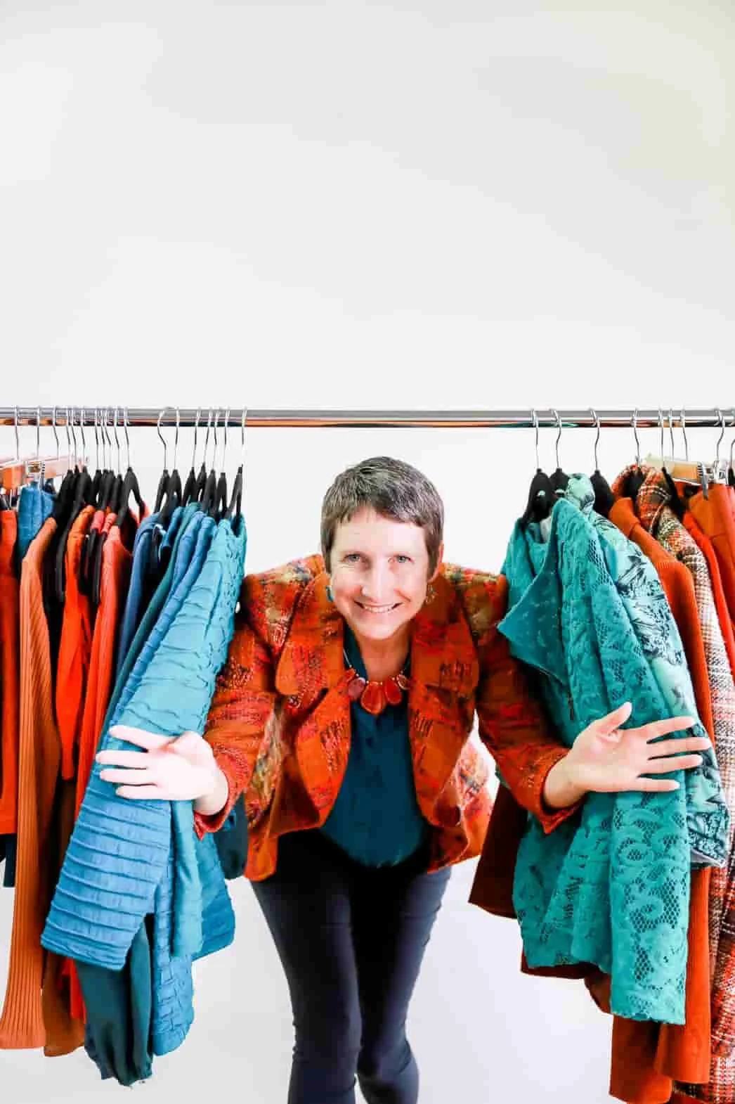 A woman stands between two clothing racks filled with colorful jackets and coats, smiling and looking at the camera.