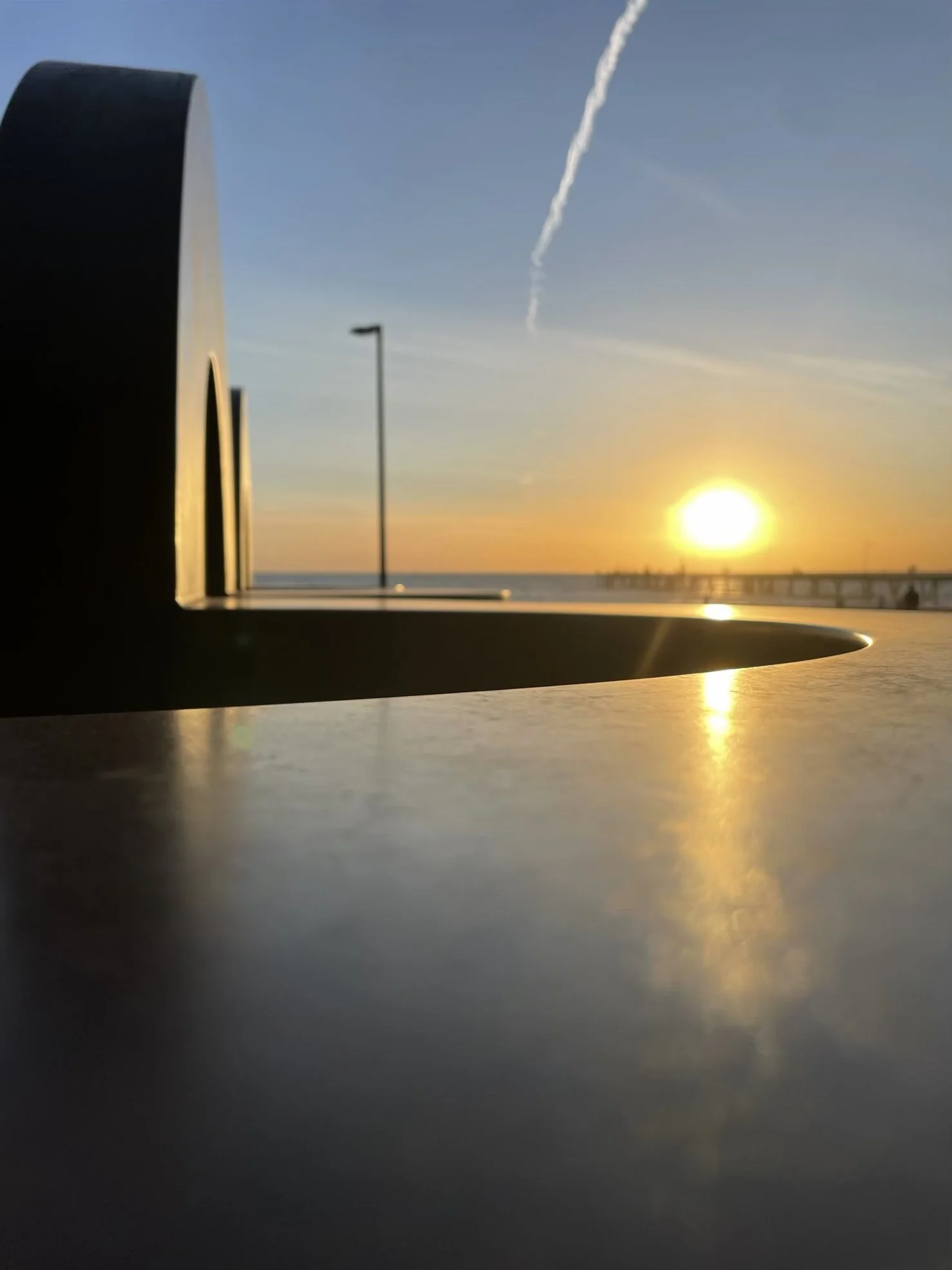 The sun setting over the Glenelg pier is reflected on the surface of a nearby sculpture.