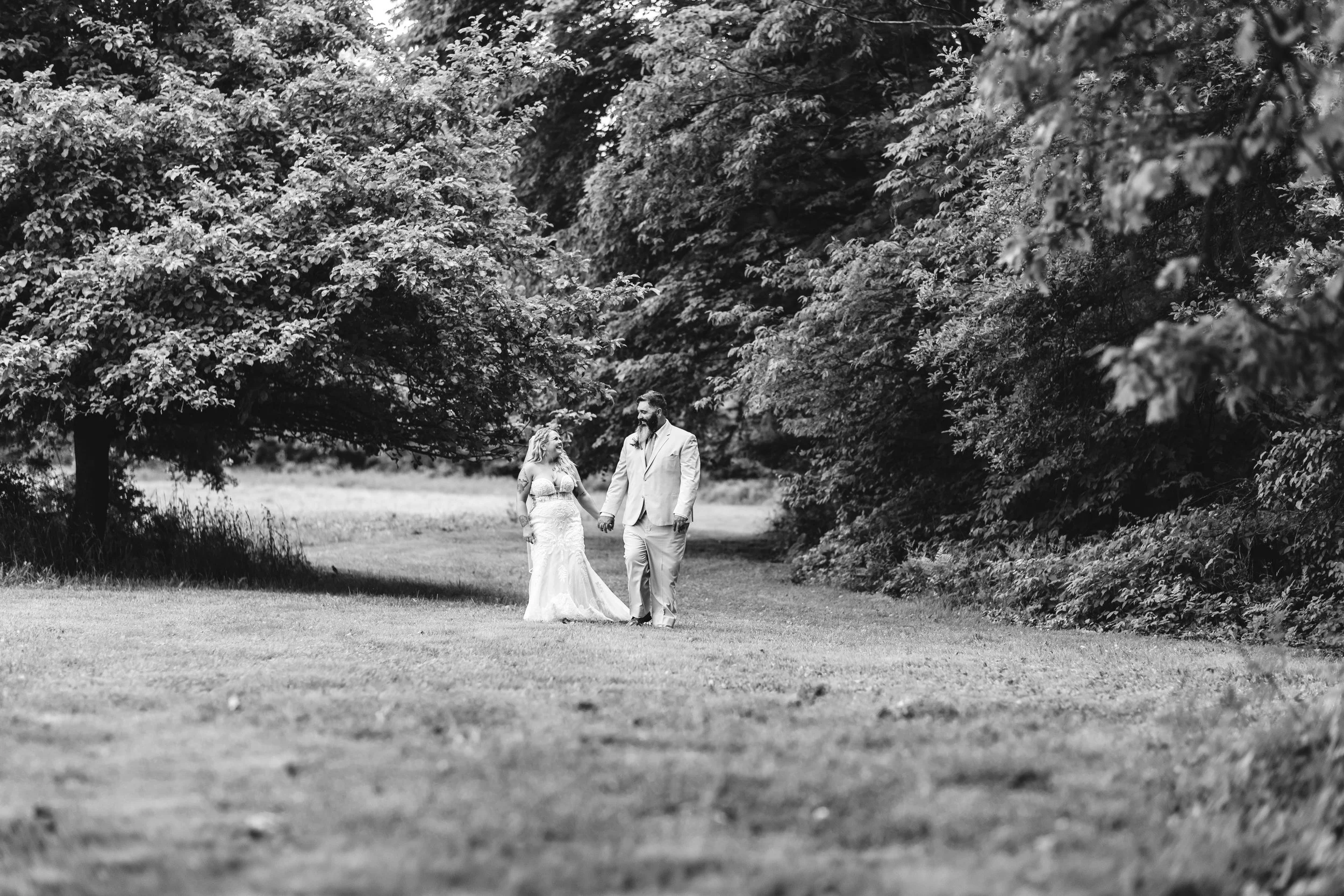 A black and white photo of a bride and groom walking hand in hand outdoors in a park surrounded by large trees.