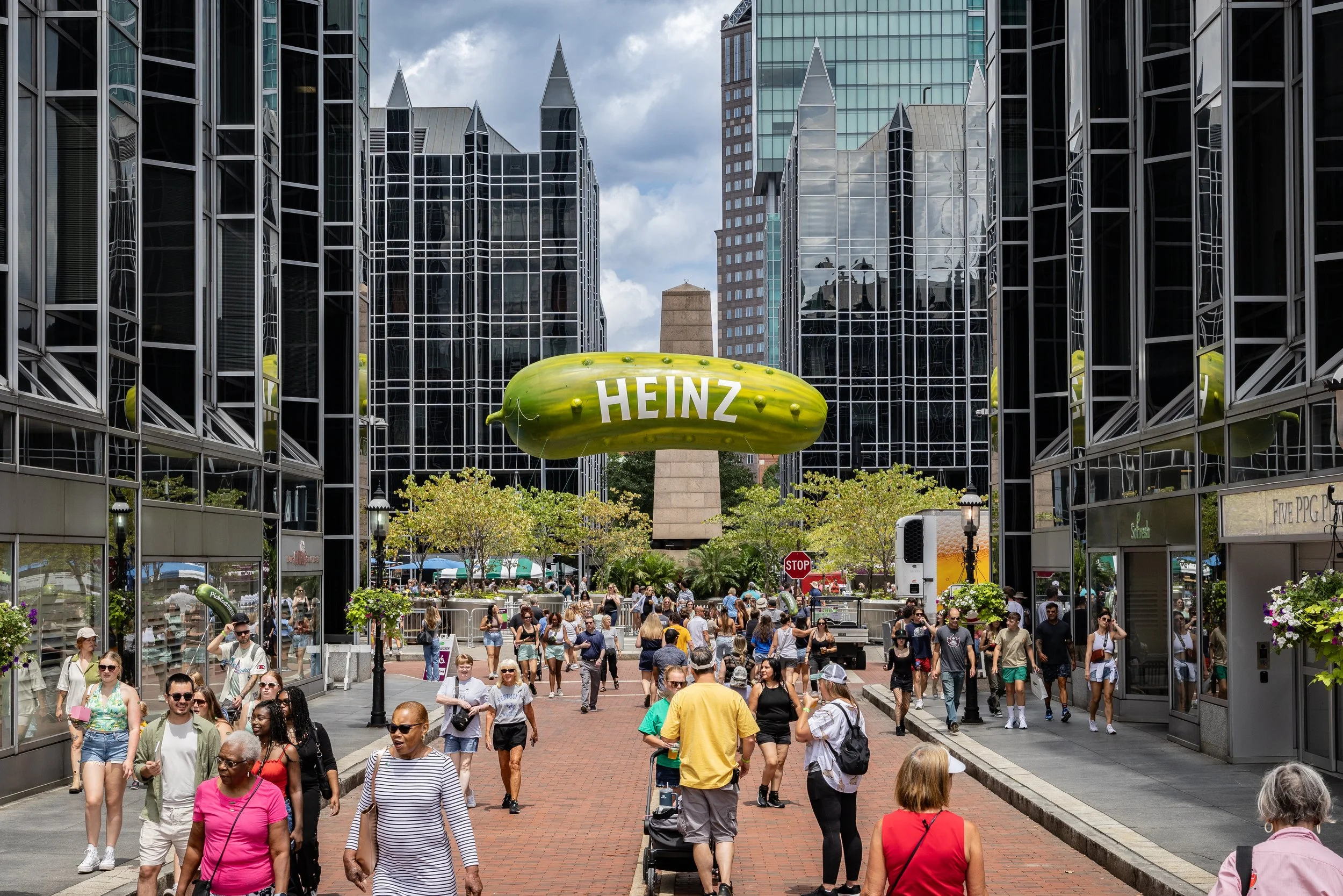 Crowd of people walking in a city street with tall glass buildings and a large Heinz condiment bottle sculpture.