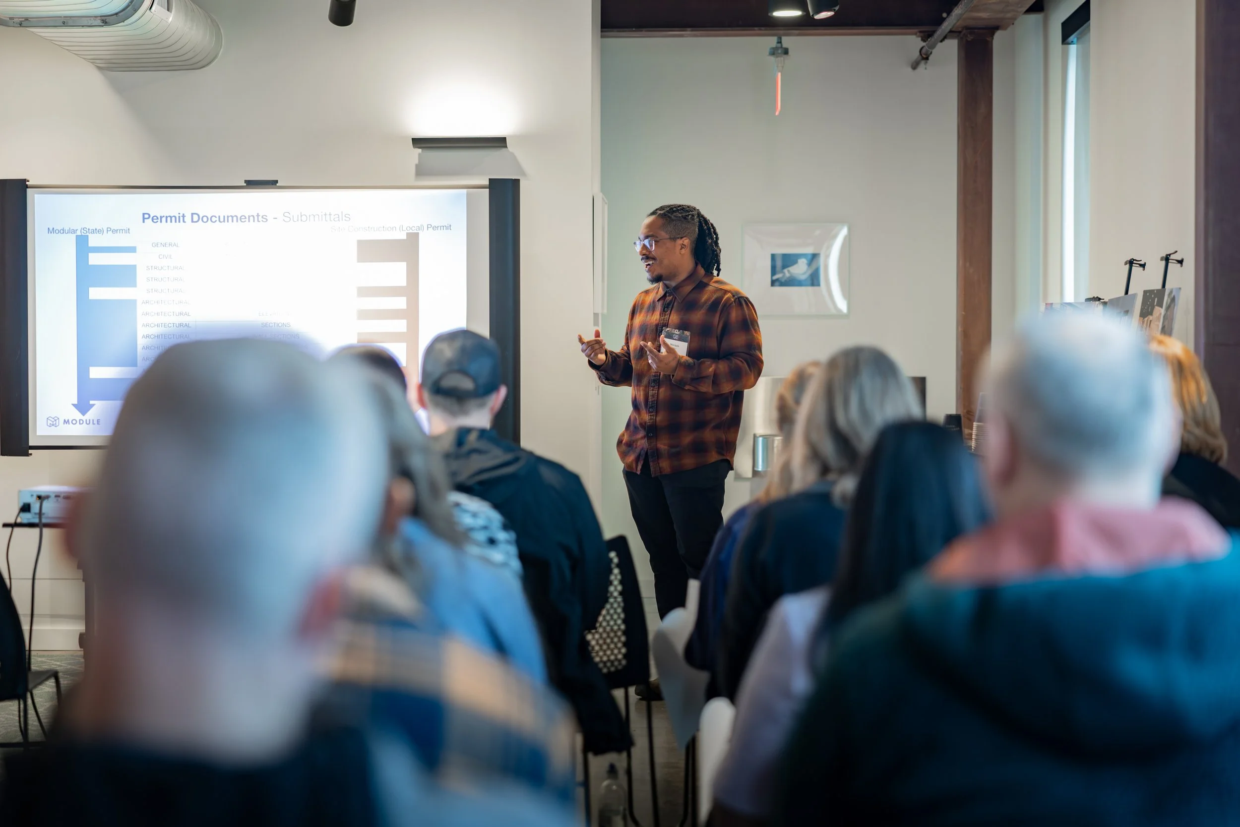 A man in a plaid shirt and glasses giving a presentation to an audience in a room with white walls and a large screen displaying a slide titled 'Permit Documents - Submittals'.