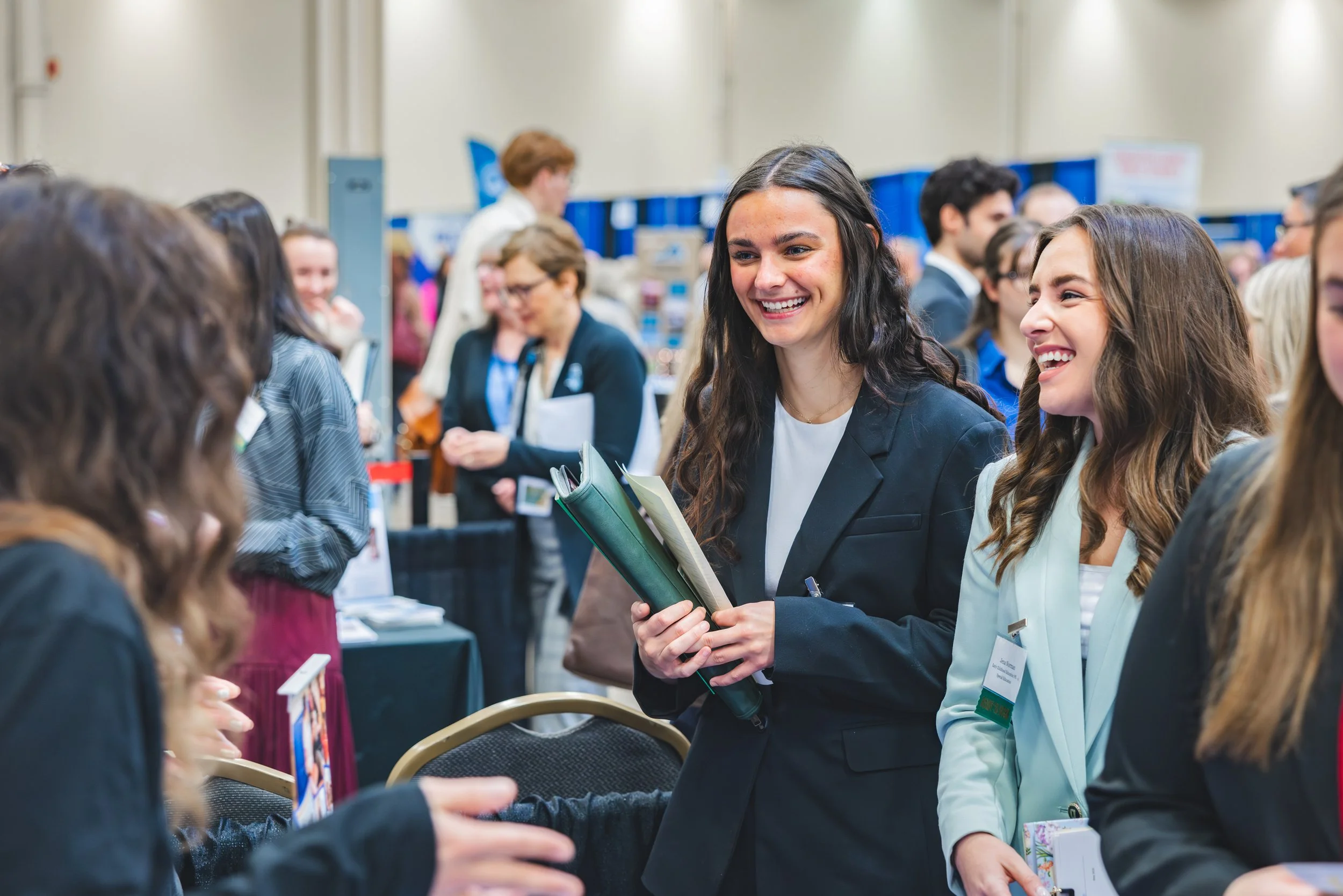 Two women in professional attire smiling and engaging in conversation at a busy conference or event, surrounded by other attendees.