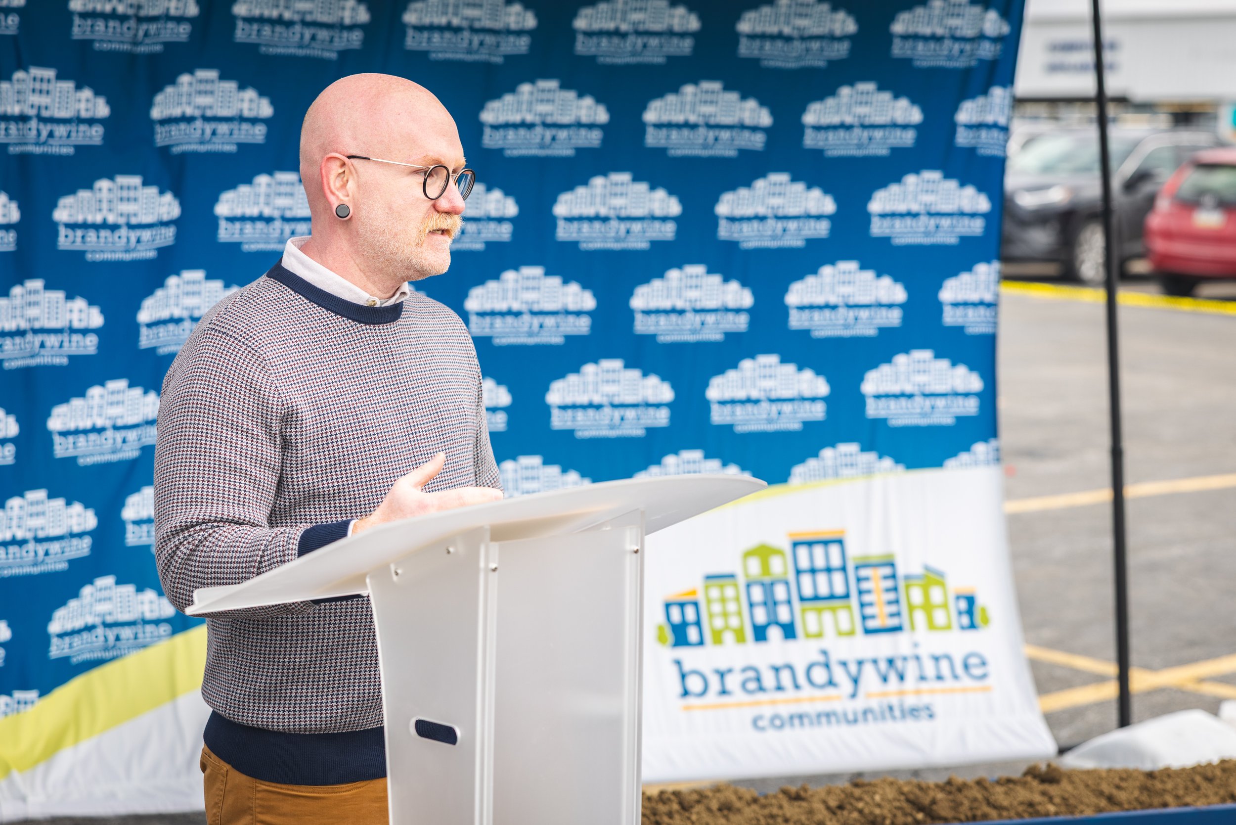 A man with glasses and earrings standing at a podium outdoors speaking at an event, with a blue branded backdrop behind him and a colorful illustrated sign in front of the podium.