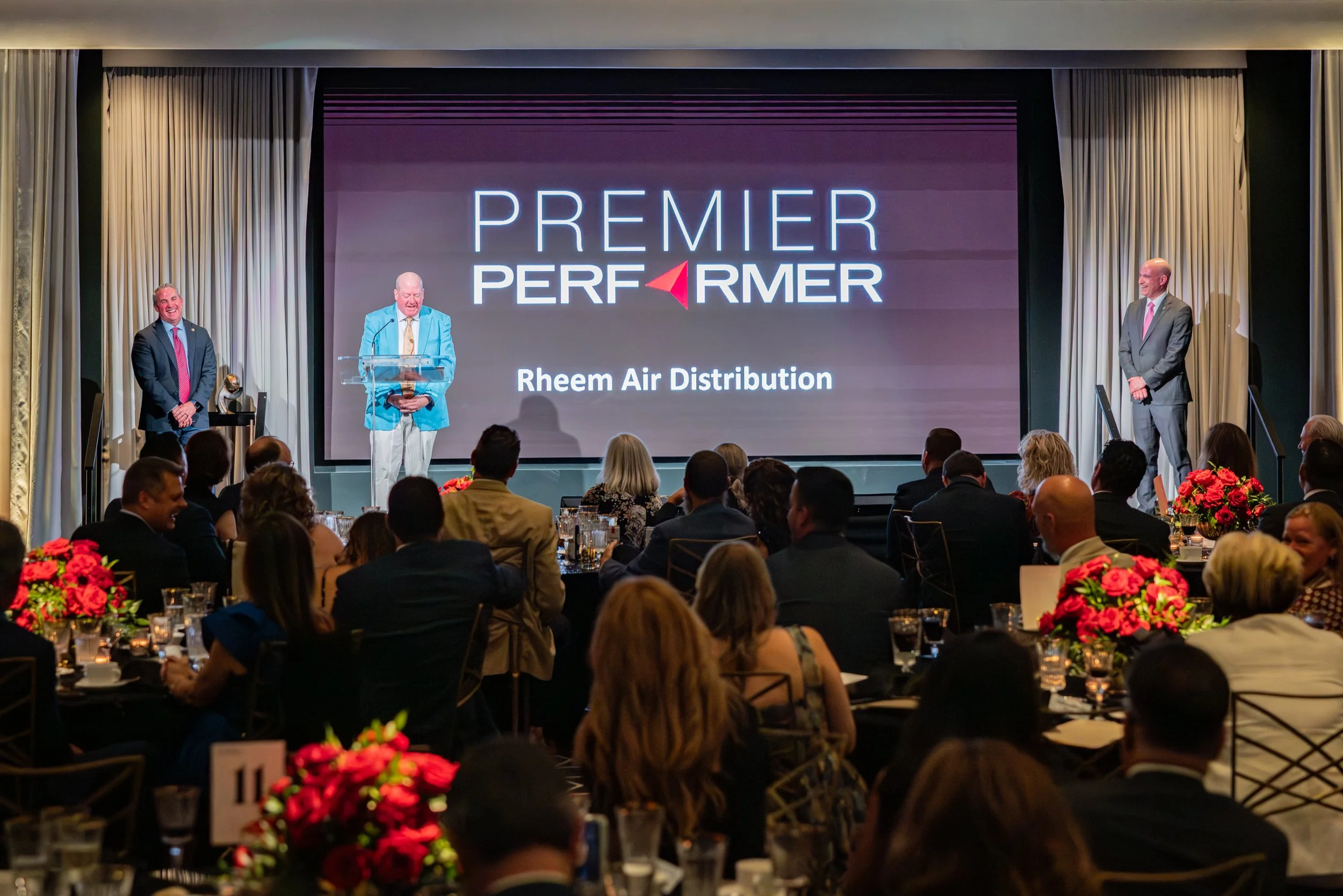 A group of people attending an awards ceremony or conference with a presentation slide that reads 'Premier Performer' and 'Rheem Air Distribution.' Three men are on stage, with one speaking at a podium. The audience is seated at tables decorated with red flowers.