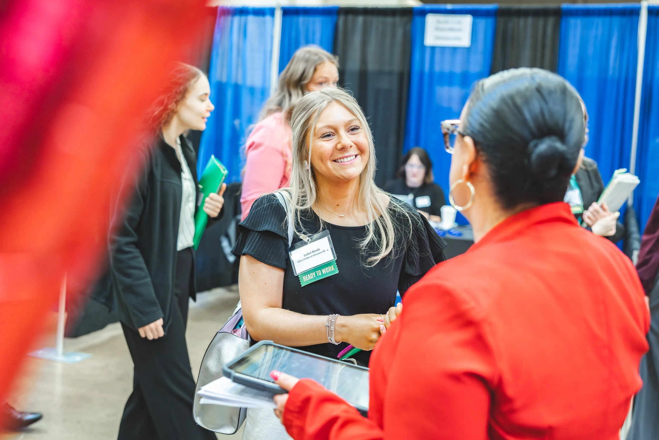 Women at an indoor event engaging in conversation, with a woman wearing a black shirt in the center smiling, holding papers, and wearing a name badge that reads "READY TO WORK." Others in the background hold documents or folders, and blue dividers are visible.