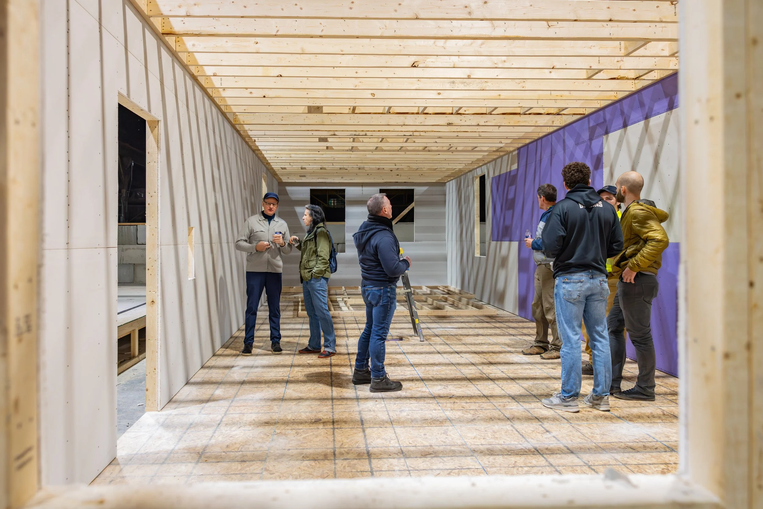 People standing and talking inside a wooden construction site with partially built walls and ceiling.
