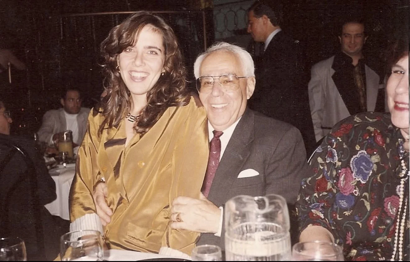 A woman with curly brown hair and a man with white hair, glasses, and a suit smiling and posing together at a social event, sitting at a table with glasses and a pitcher in front of them, surrounded by others in formal attire.