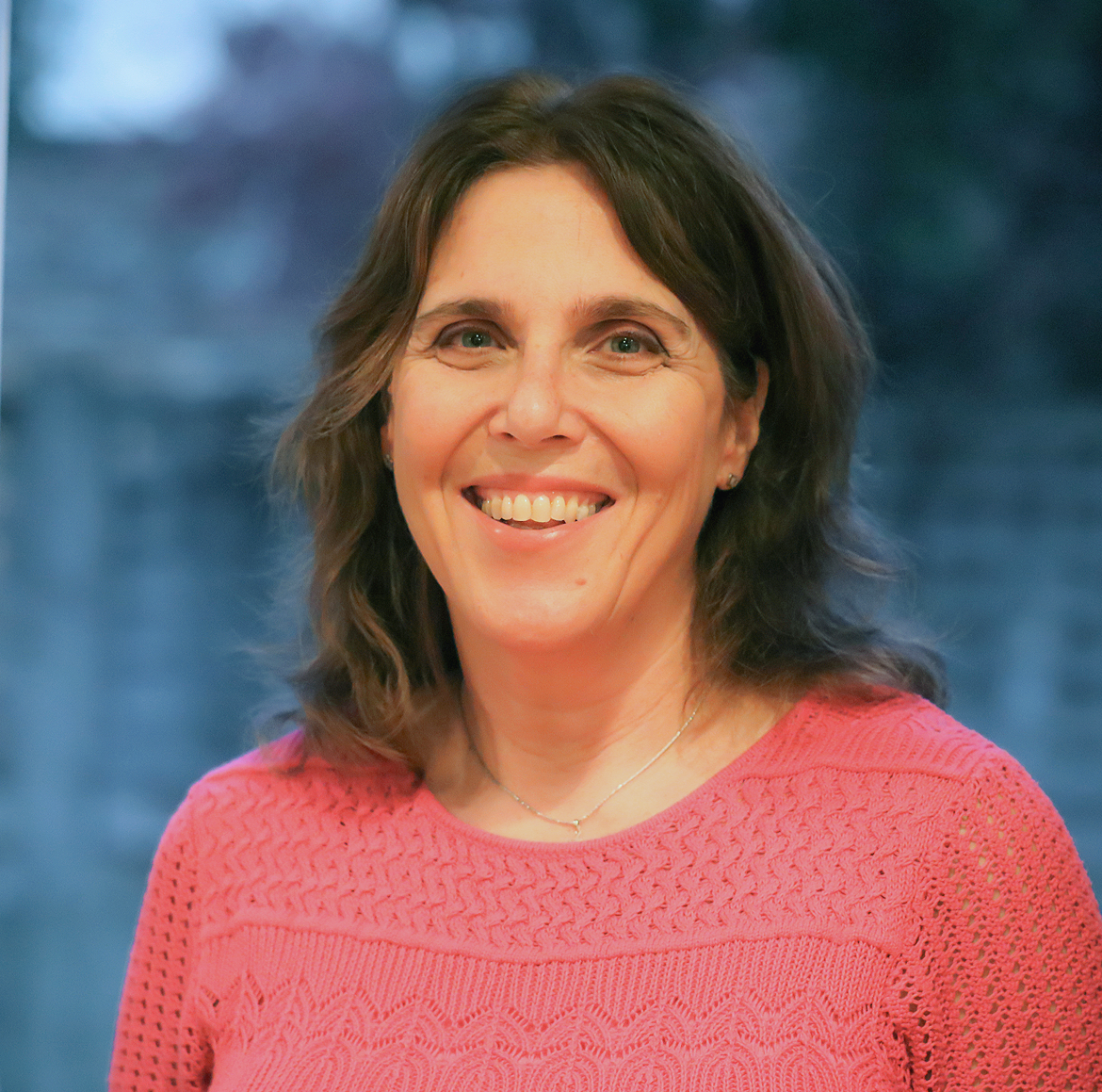 A woman with shoulder-length dark brown hair, smiling, wearing a pink knitted sweater and a delicate necklace, standing outdoors with a blurred blue sky and trees in the background.