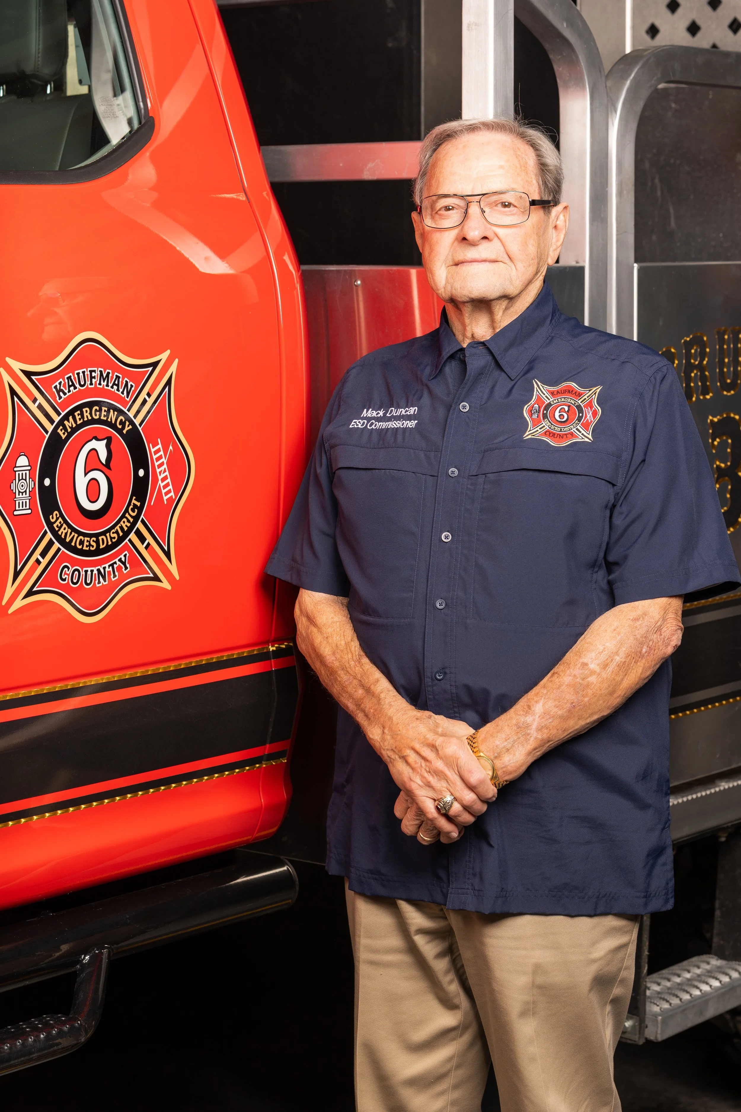 An elderly man with glasses, gray hair, and wearing a light blue collared shirt in front of a plain white background.