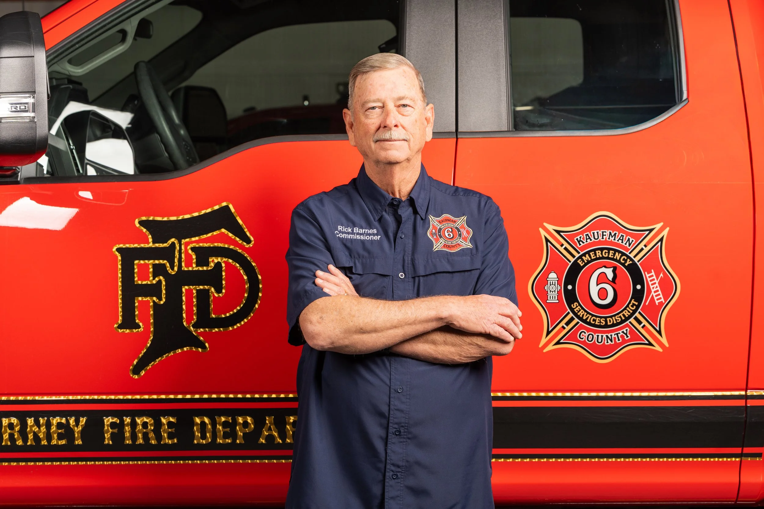 A smiling elderly man with a mustache, wearing a black collared shirt, standing against a plain light-colored background.