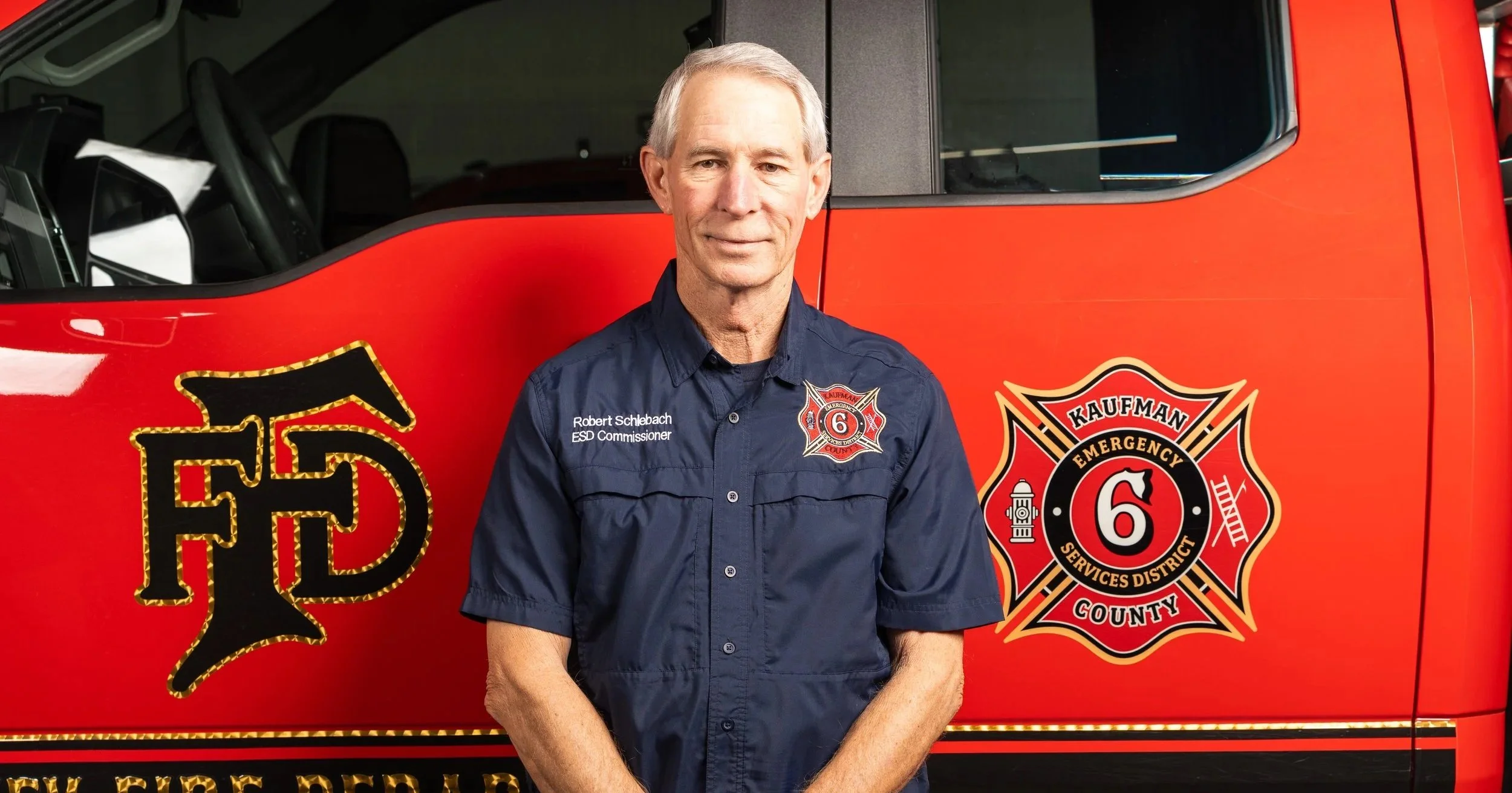 Close-up portrait of a middle-aged man with gray hair and light skin, wearing a plaid collared shirt, smiling gently against a neutral background.