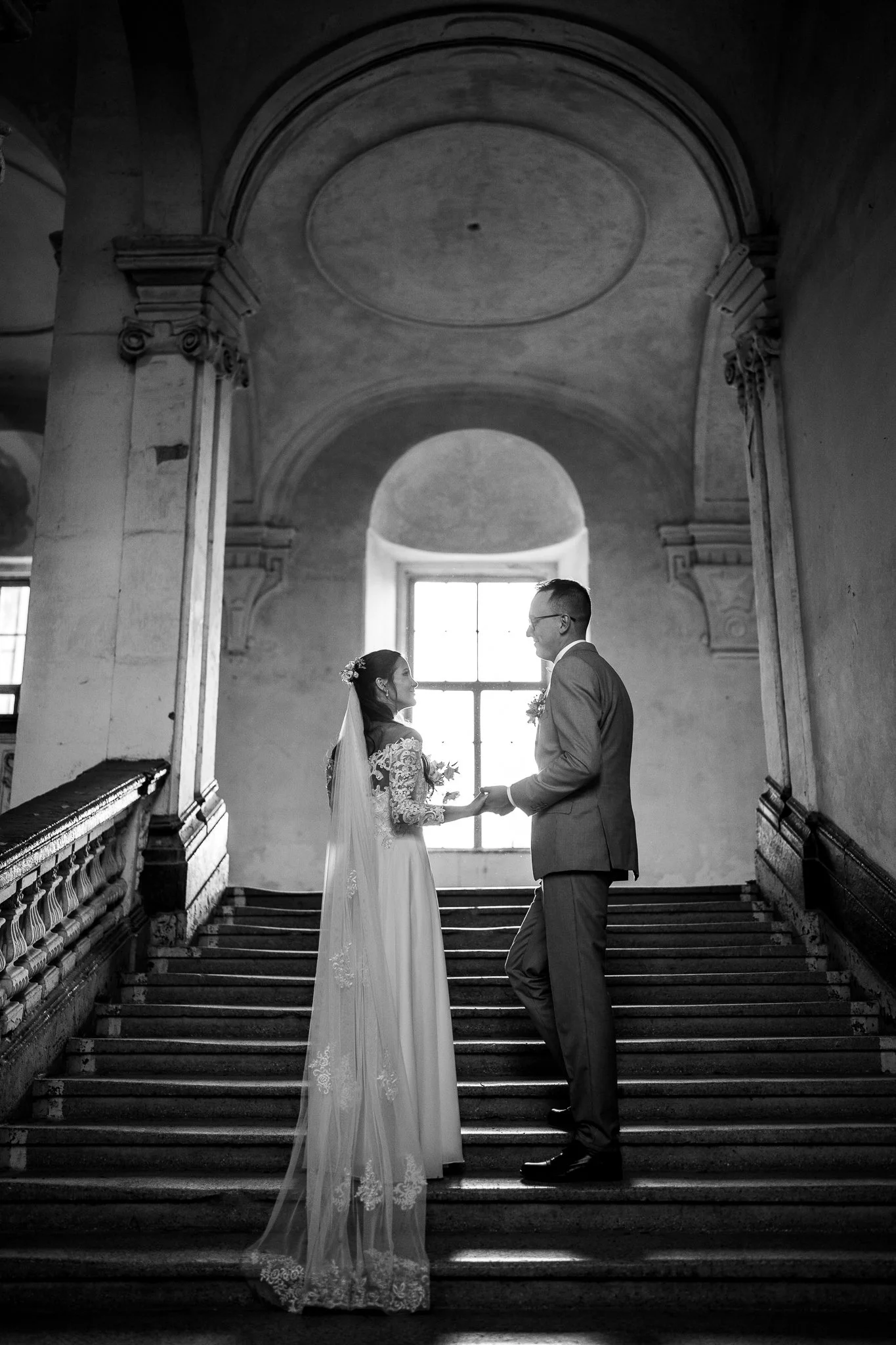 Černobílá svatební fotografie na schodišti v historické budově během svatebního dne. 