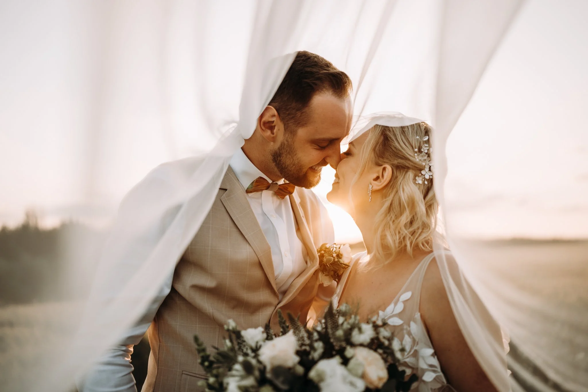 Novomanželé se na sebe smějí a mají přes sebe přehozený závoj. Krásná fotografie při západu slunce se závojem a šťastným párem.