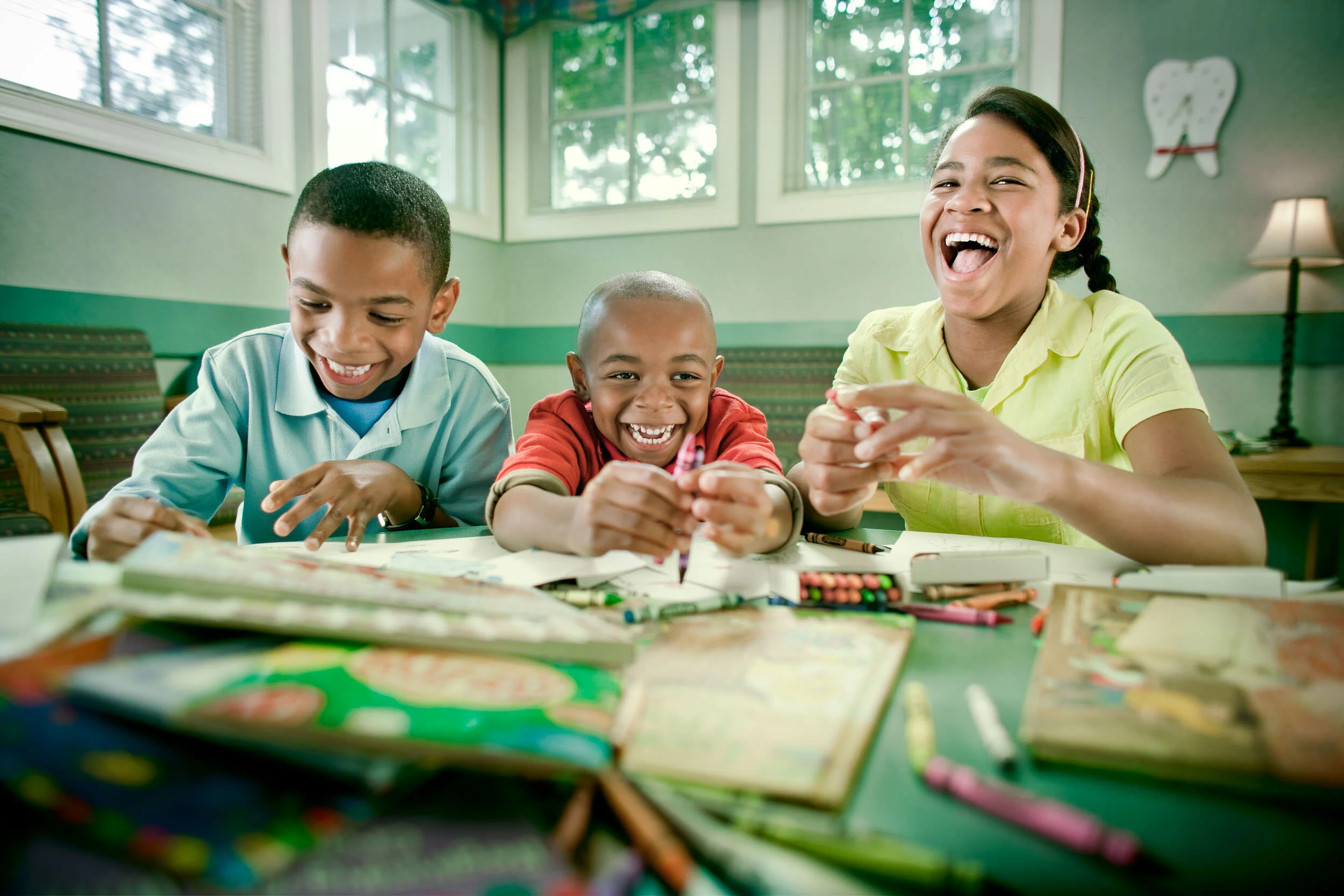 Three children sitting at a table laughing and coloring with crayons in a bright room.