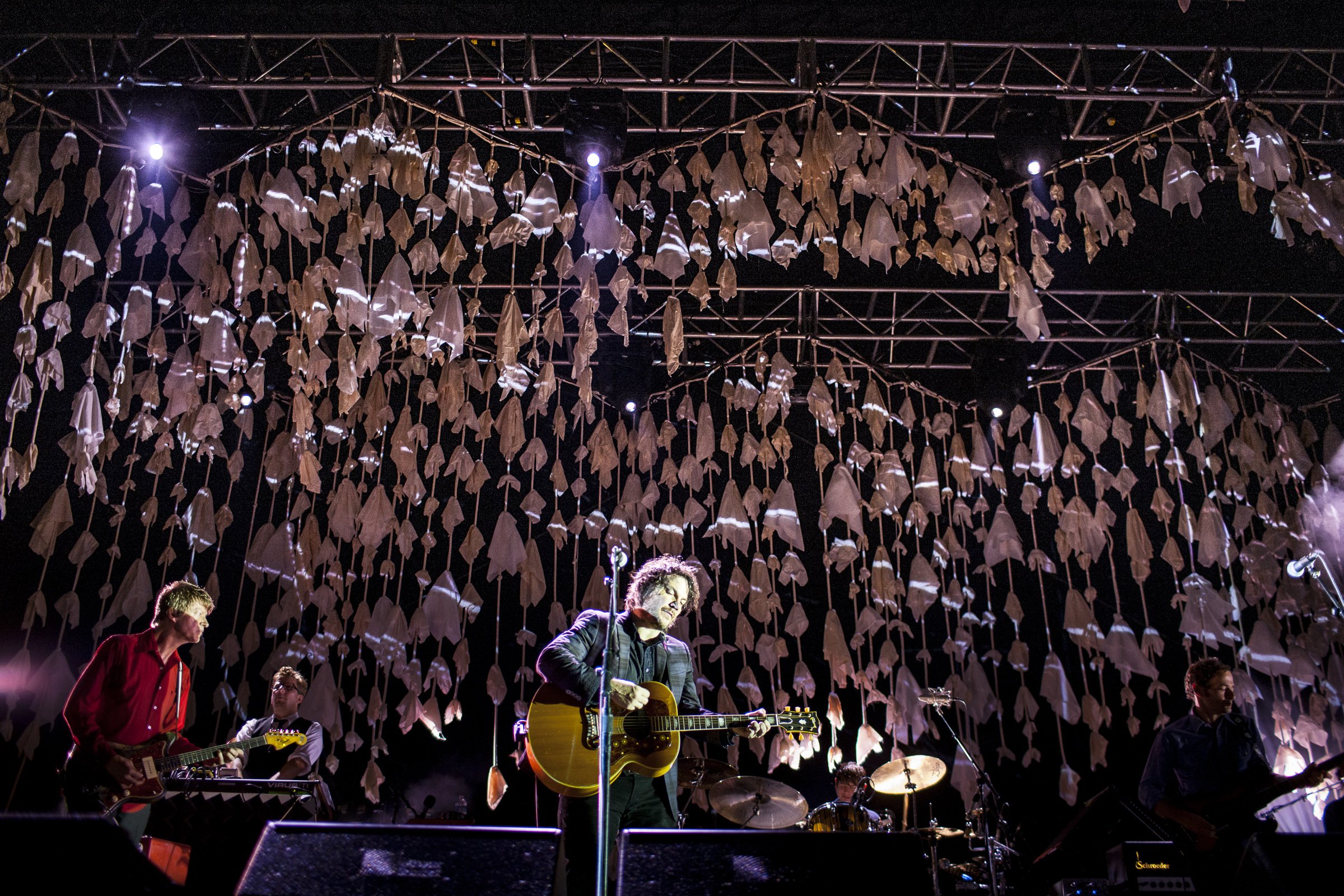 A band performing on stage with a backdrop of hanging white fabric decorations, illuminated by stage lights.